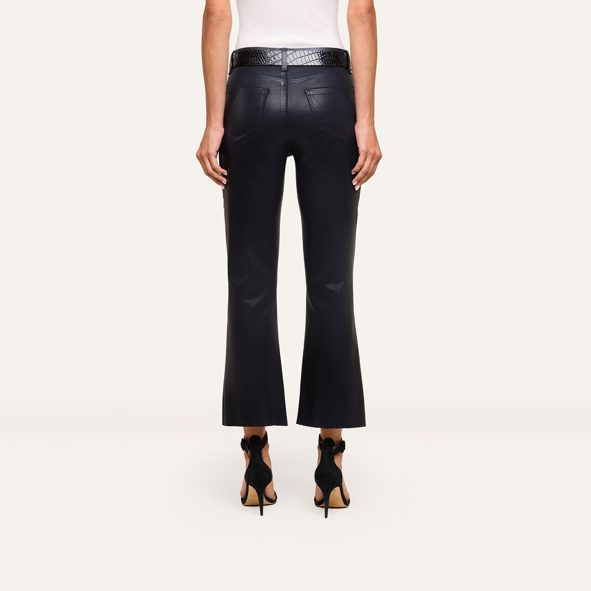 Someone wearing the Kendra Coated Cropped Flare Jean, a white sleeveless top, black heels, and a textured black belt stands with their back to the camera against a plain background.