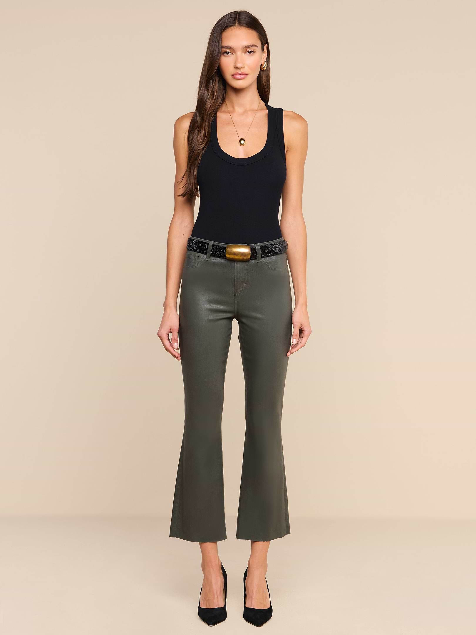 A woman stands against a plain white background, wearing the Kendra Coated Cropped Flare Jean in ash olive green, paired with a black tank top, black belt, and black heels. She has long brown hair and is looking directly at the camera.