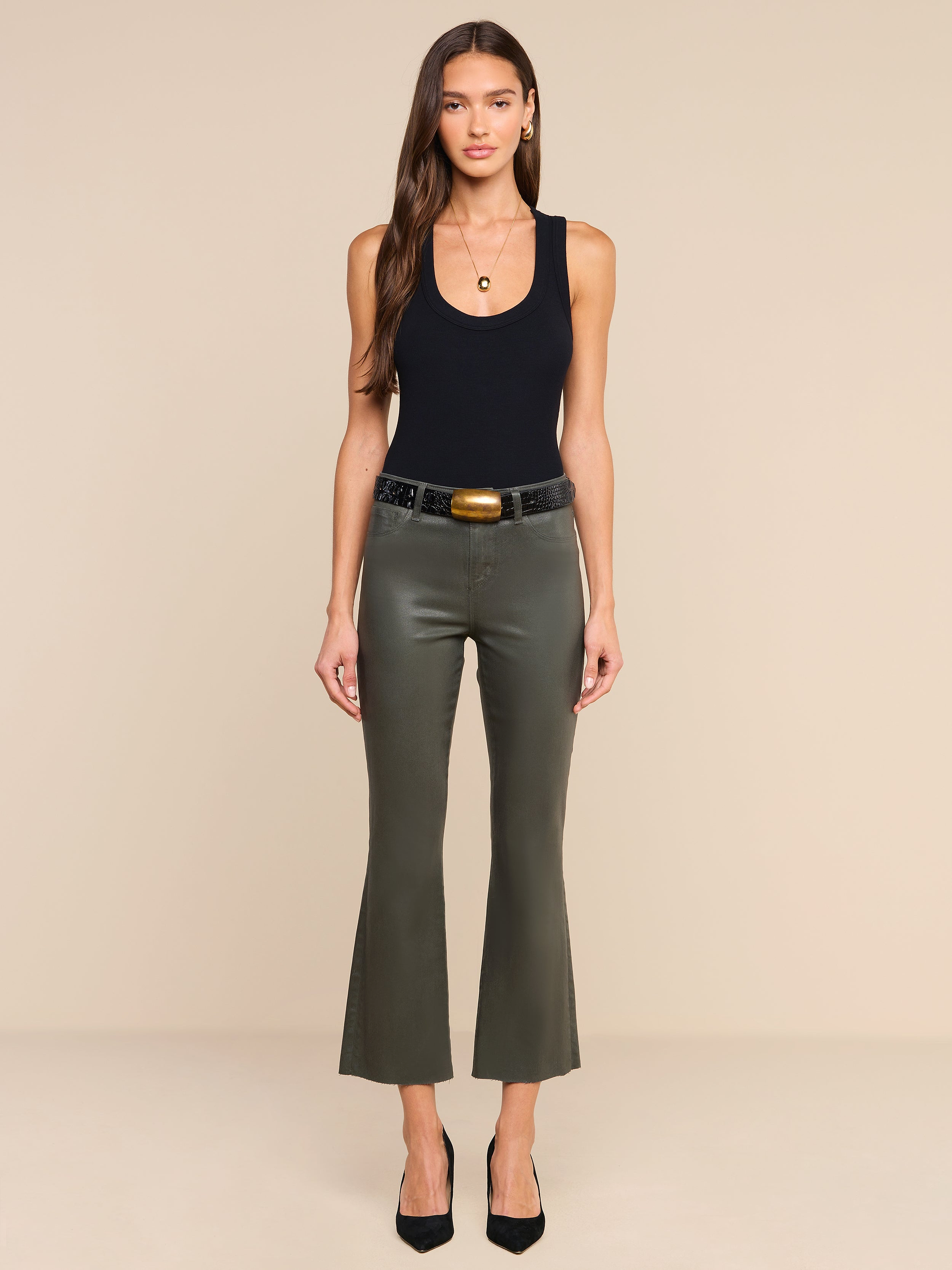 A woman stands facing forward, wearing the Kendra Coated Cropped Flare Jean in ash olive green with a black sleeveless top, a black belt featuring a large buckle, and black heels. She has long brown hair and minimal jewelry, against a plain beige background.