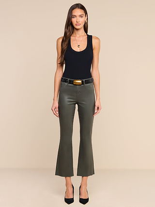 A woman stands against a plain white background, wearing the Kendra Coated Cropped Flare Jean in ash olive green, paired with a black tank top, black belt, and black heels. She has long brown hair and is looking directly at the camera.