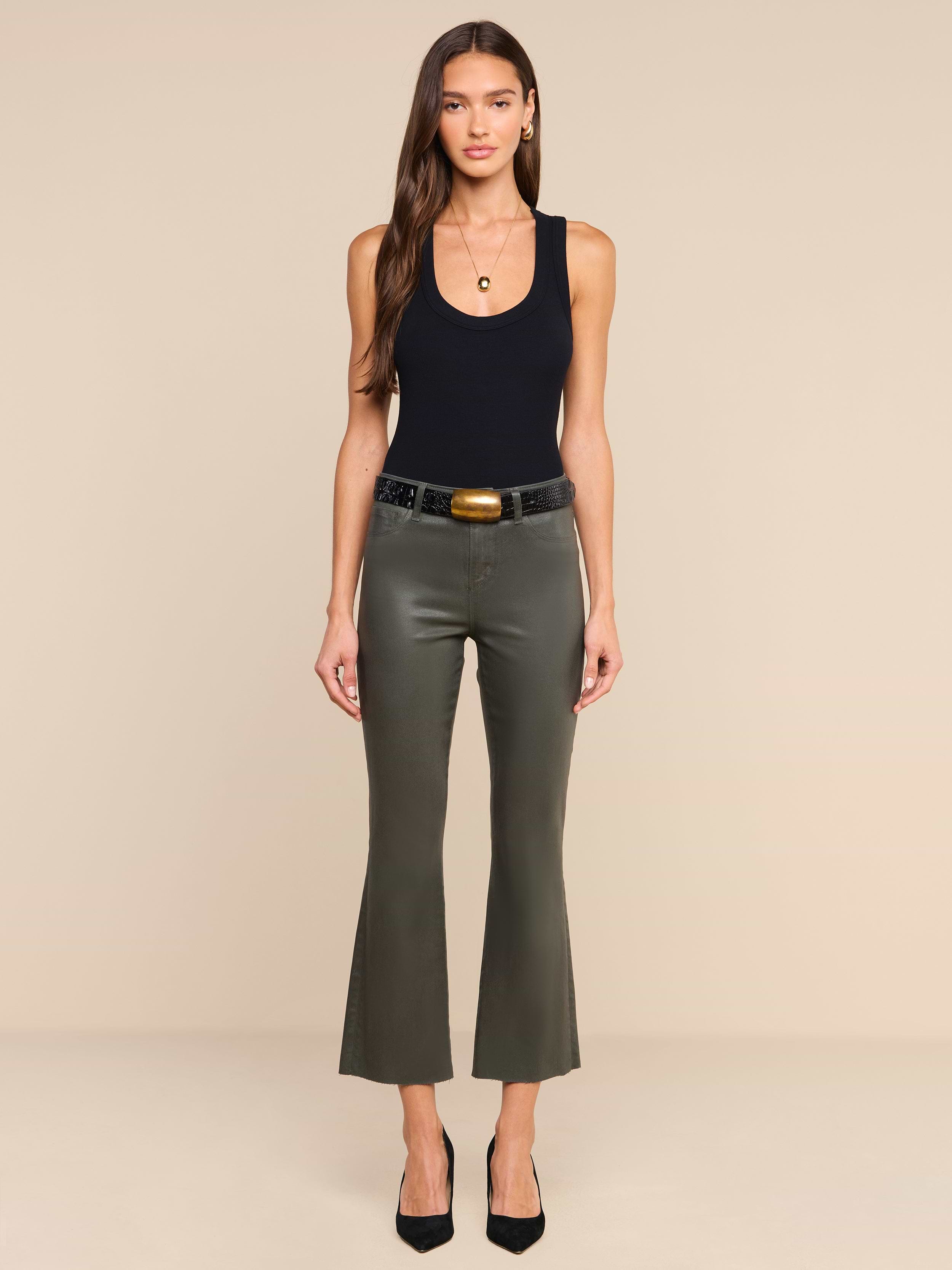 A woman stands facing forward, wearing the Kendra Coated Cropped Flare Jean in ash olive green with a black sleeveless top, a black belt featuring a large buckle, and black heels. She has long brown hair and minimal jewelry, against a plain beige background.