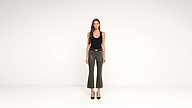 A woman with long brown hair stands facing forward, wearing a black tank top, olive green flared pants, and black heels, against a plain white background.