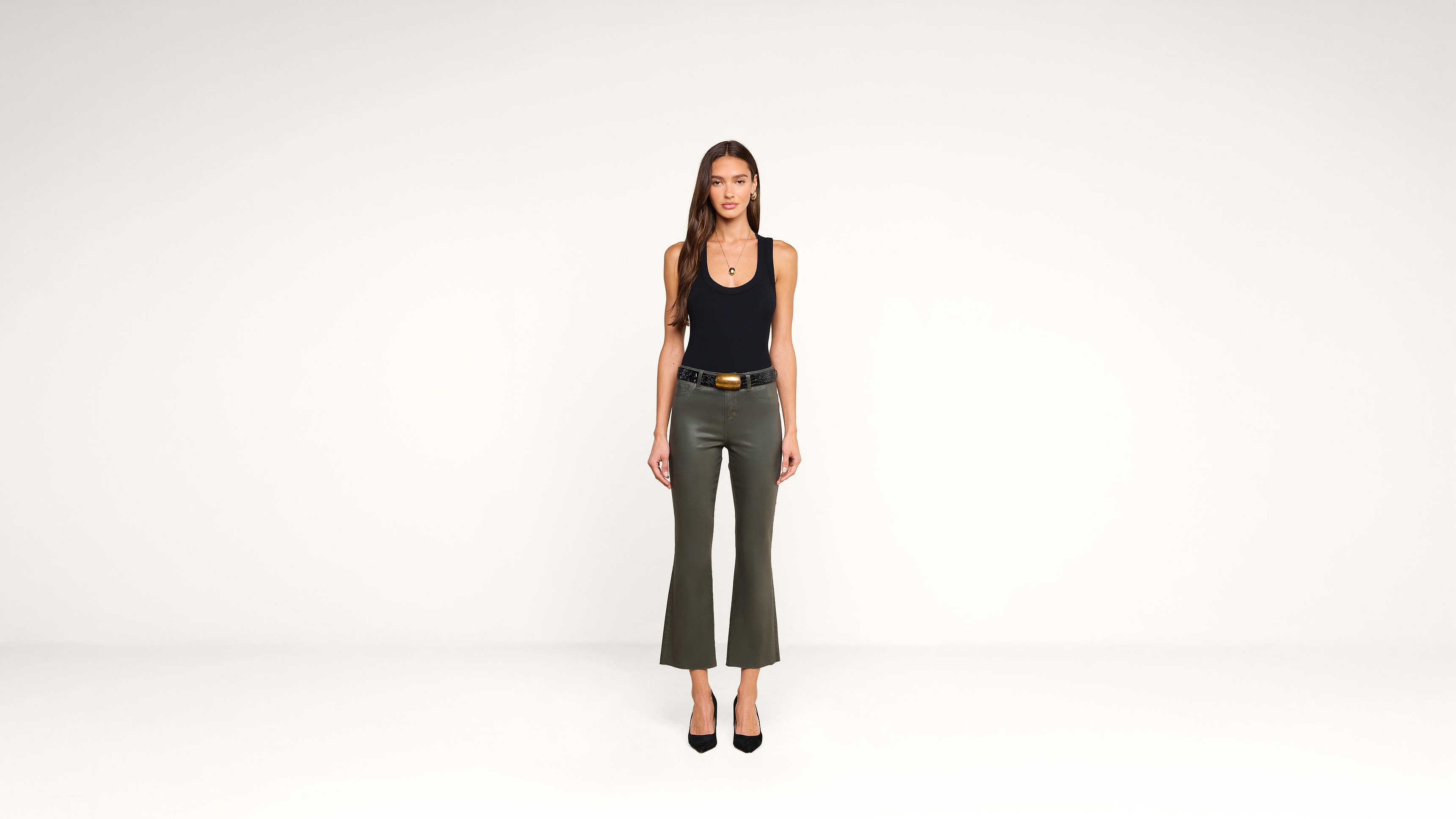A woman with long brown hair stands facing forward, wearing a black tank top, olive green flared pants, and black heels, against a plain white background.