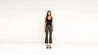 A woman stands against a plain white background, wearing a black sleeveless top, gray-green pants, black heels, and a belt. She faces forward with her arms relaxed at her sides, showcasing a modern and minimalist outfit.