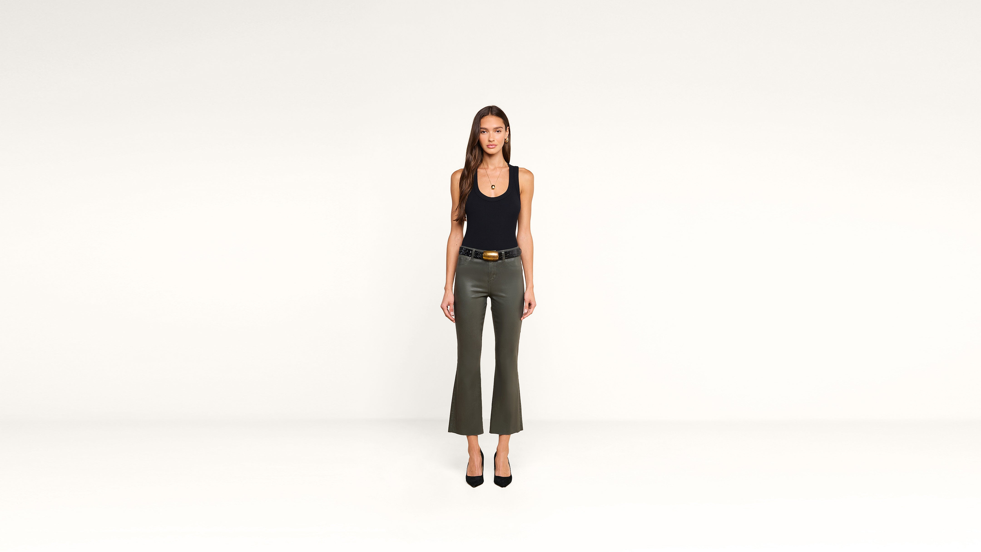 A woman stands against a plain white background, wearing a black sleeveless top, gray-green pants, black heels, and a belt. She faces forward with her arms relaxed at her sides, showcasing a modern and minimalist outfit.