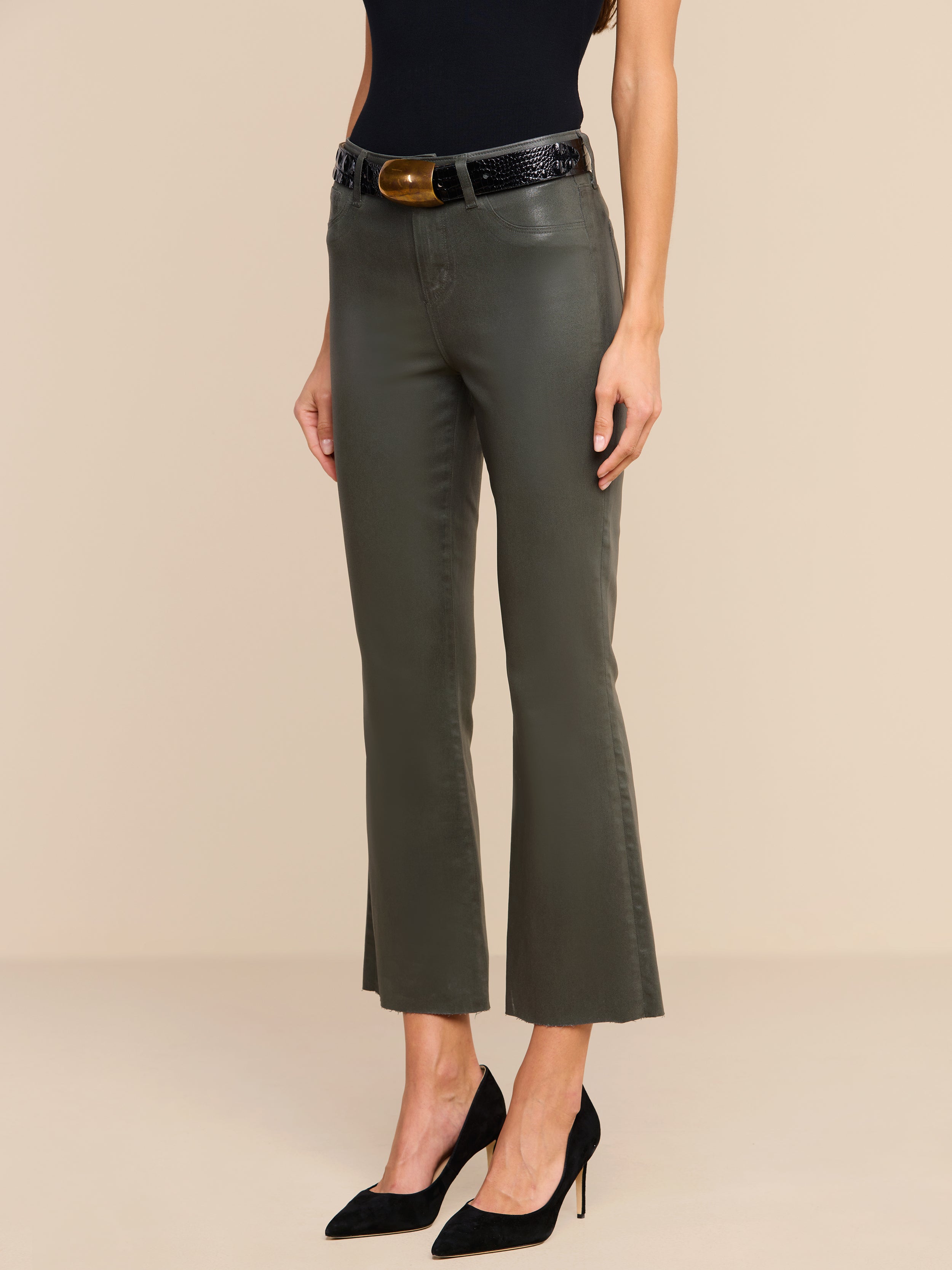 Woman wearing Kendra Coated Cropped Flare Jeans in ash olive green, a black sleeveless top, a black belt with a large buckle, and black pointed high heels, standing against a plain beige background.