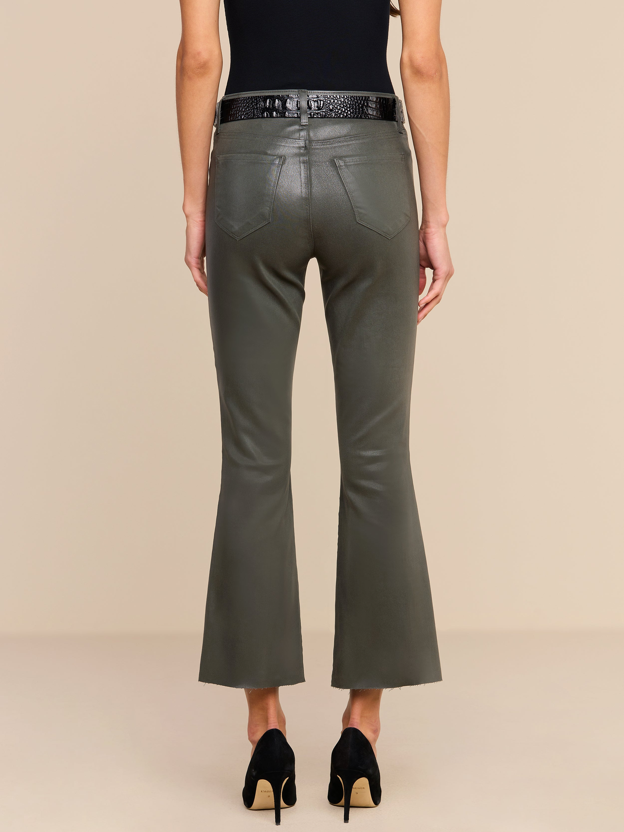A person wearing the Kendra Coated Cropped Flare Jean in ash olive green, styled with a black belt, black sleeveless top, and black heels, is shown from the back against a beige background.