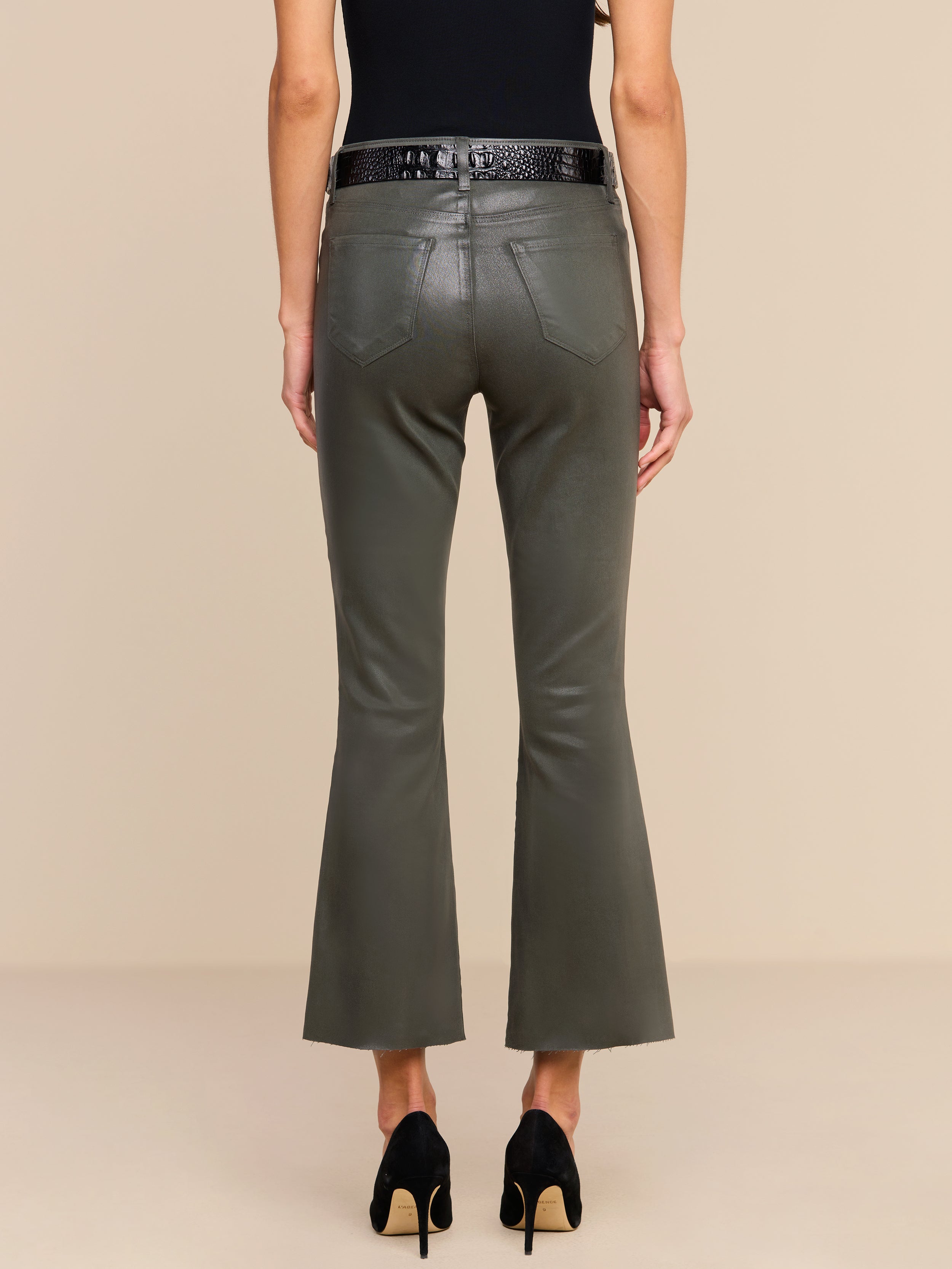 A person wearing the Kendra Coated Cropped Flare Jean in ash olive green, styled with a black belt, black sleeveless top, and black heels, is shown from the back against a beige background.