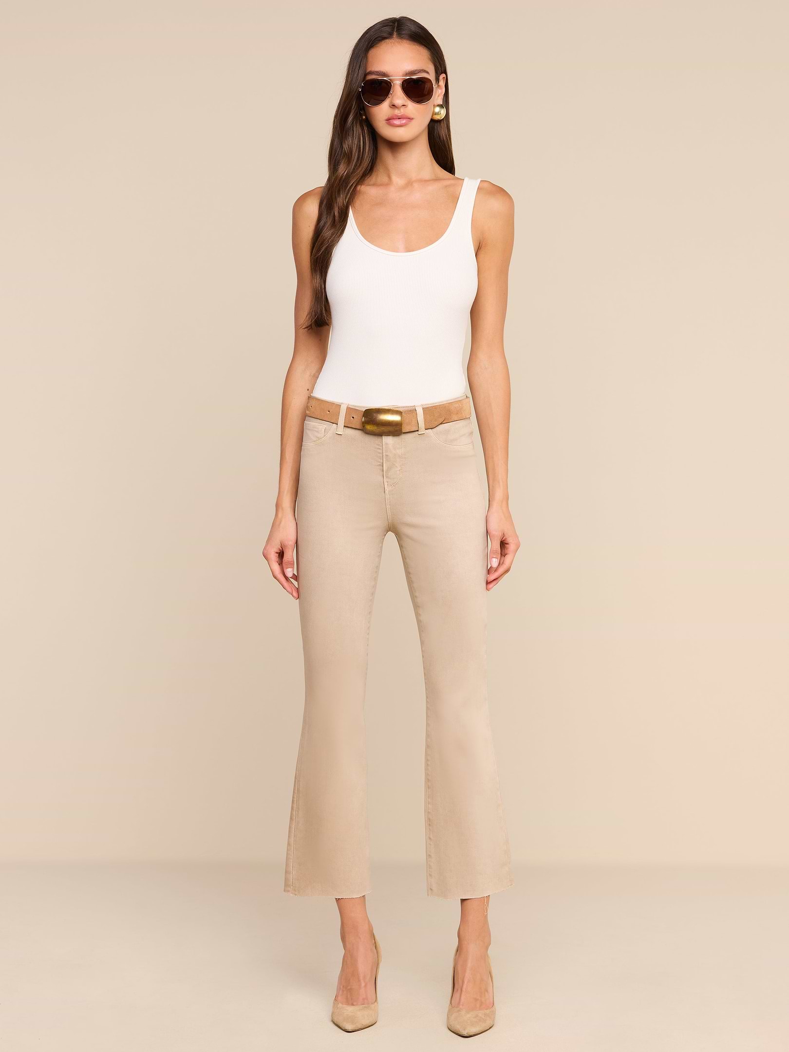 A woman stands against a plain white background, facing forward with her hands relaxed by her sides, wearing sunglasses, a white tank top, beige heels, and the Kendra Coated Cropped Flare Jean in high-rise beige.