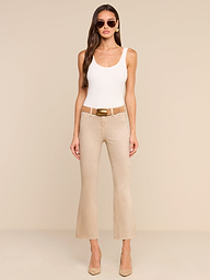 A woman stands against a plain white background, facing forward with her hands relaxed by her sides, wearing sunglasses, a white tank top, beige heels, and the Kendra Coated Cropped Flare Jean in high-rise beige.