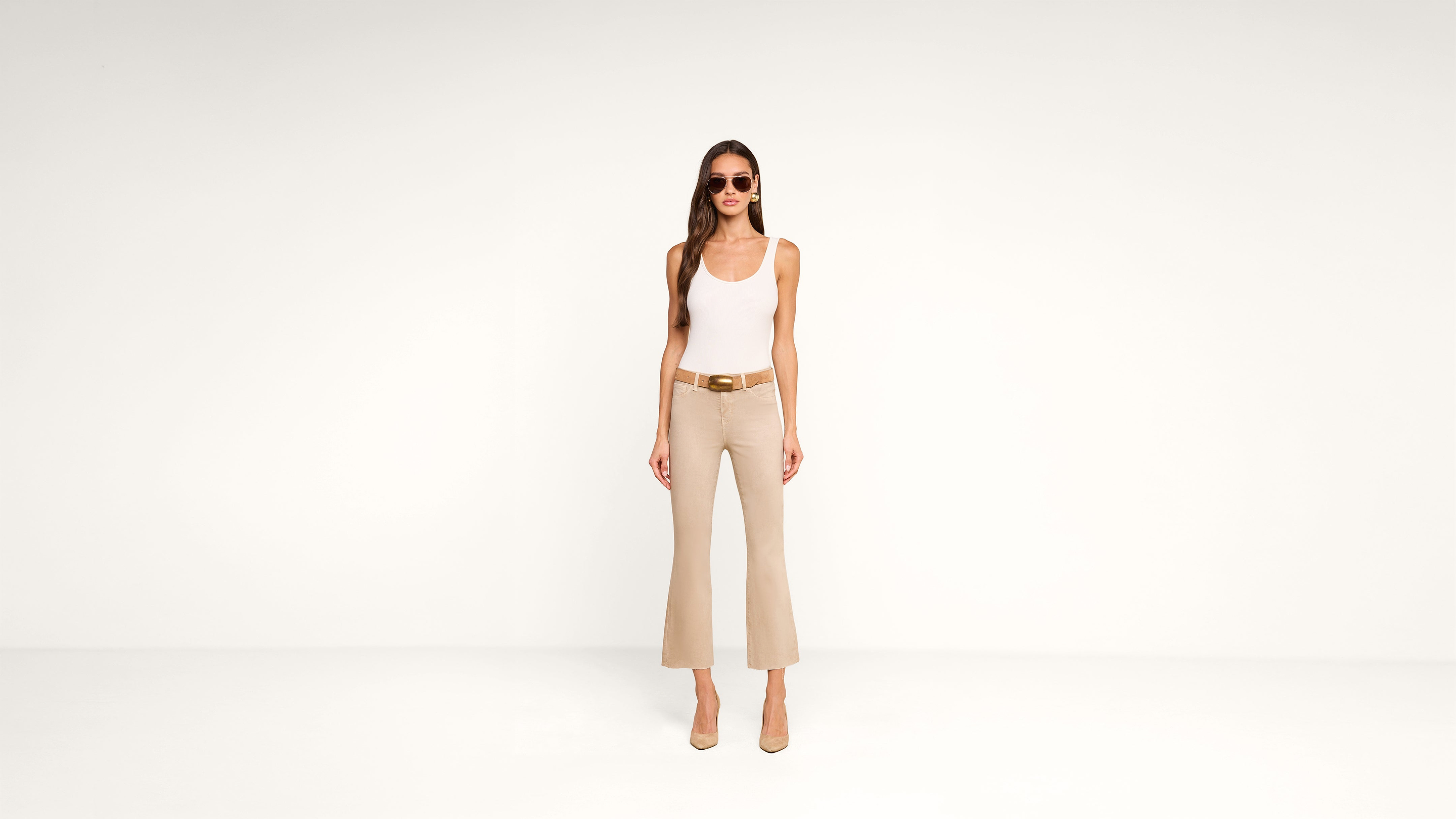 Woman stands against a plain white background wearing sunglasses, a white tank top, beige flared pants, a brown belt, and beige heels.