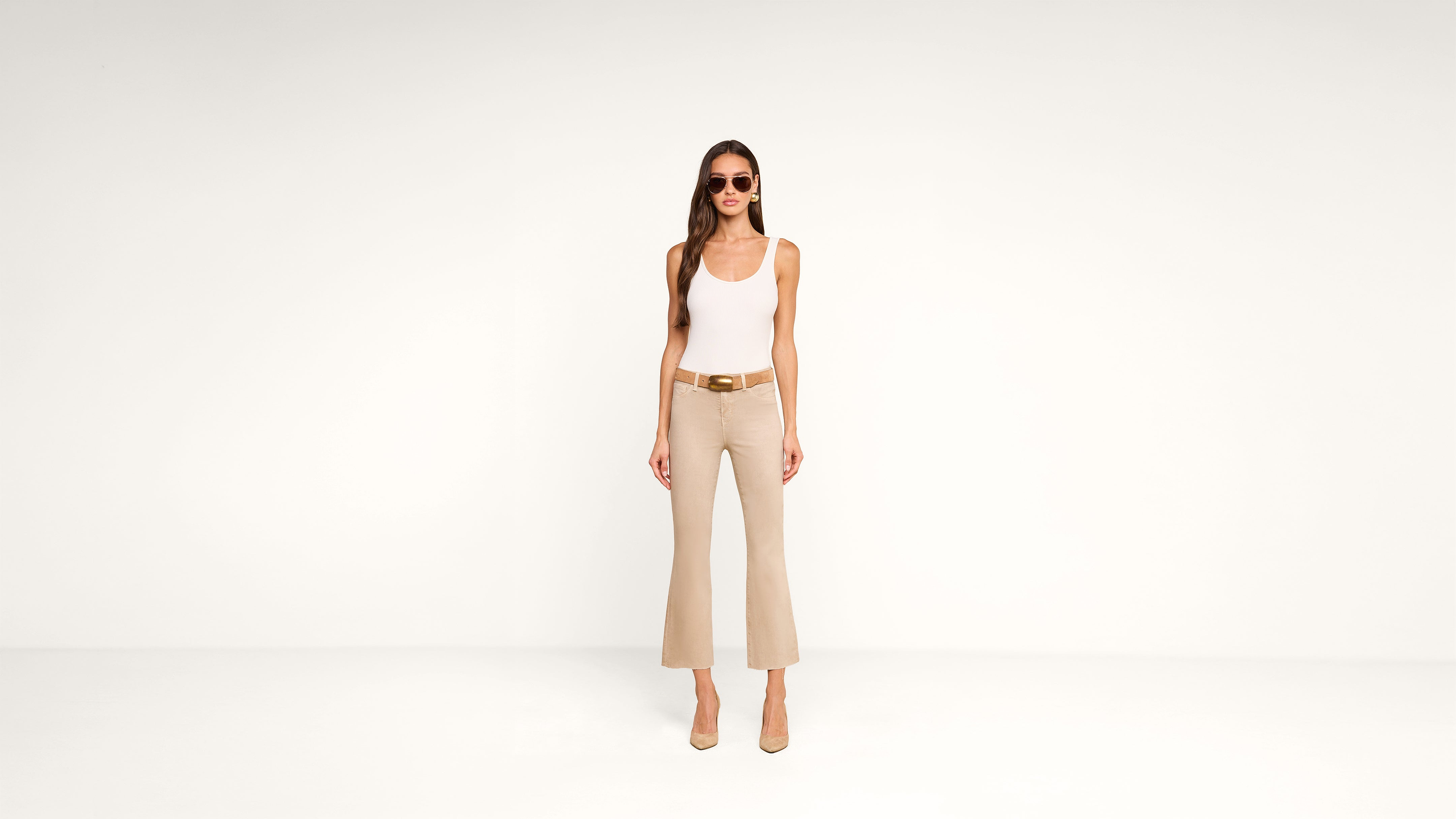Woman stands against a plain white background wearing sunglasses, a white tank top, beige flared pants, a brown belt, and beige heels.