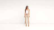 A woman wearing sunglasses, a white tank top, beige flared pants, and heels stands against a plain white background, facing forward with her hands by her sides in a chic, minimalist style.