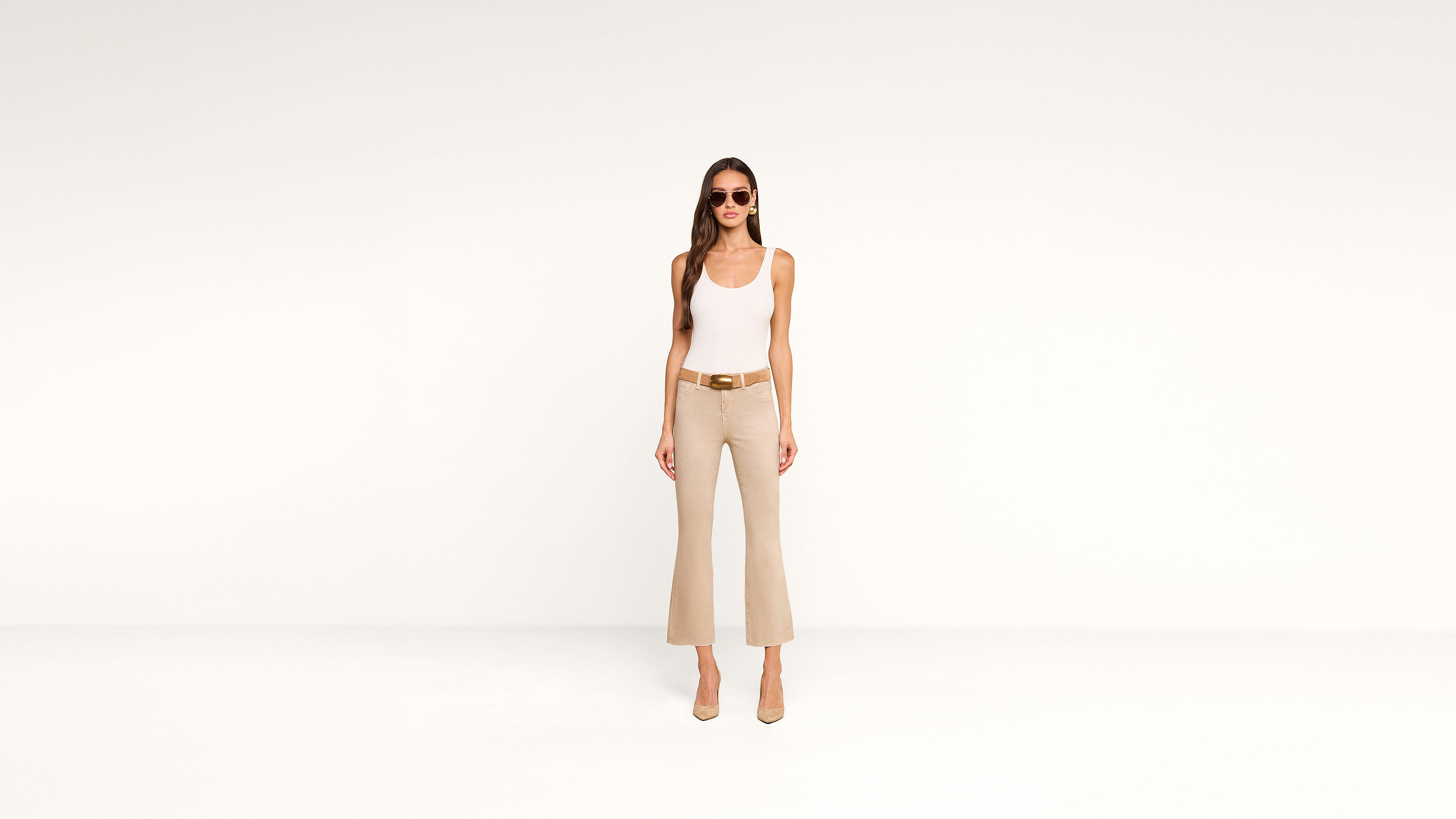 A woman wearing sunglasses, a white tank top, beige flared pants, and heels stands against a plain white background, facing forward with her hands by her sides in a chic, minimalist style.