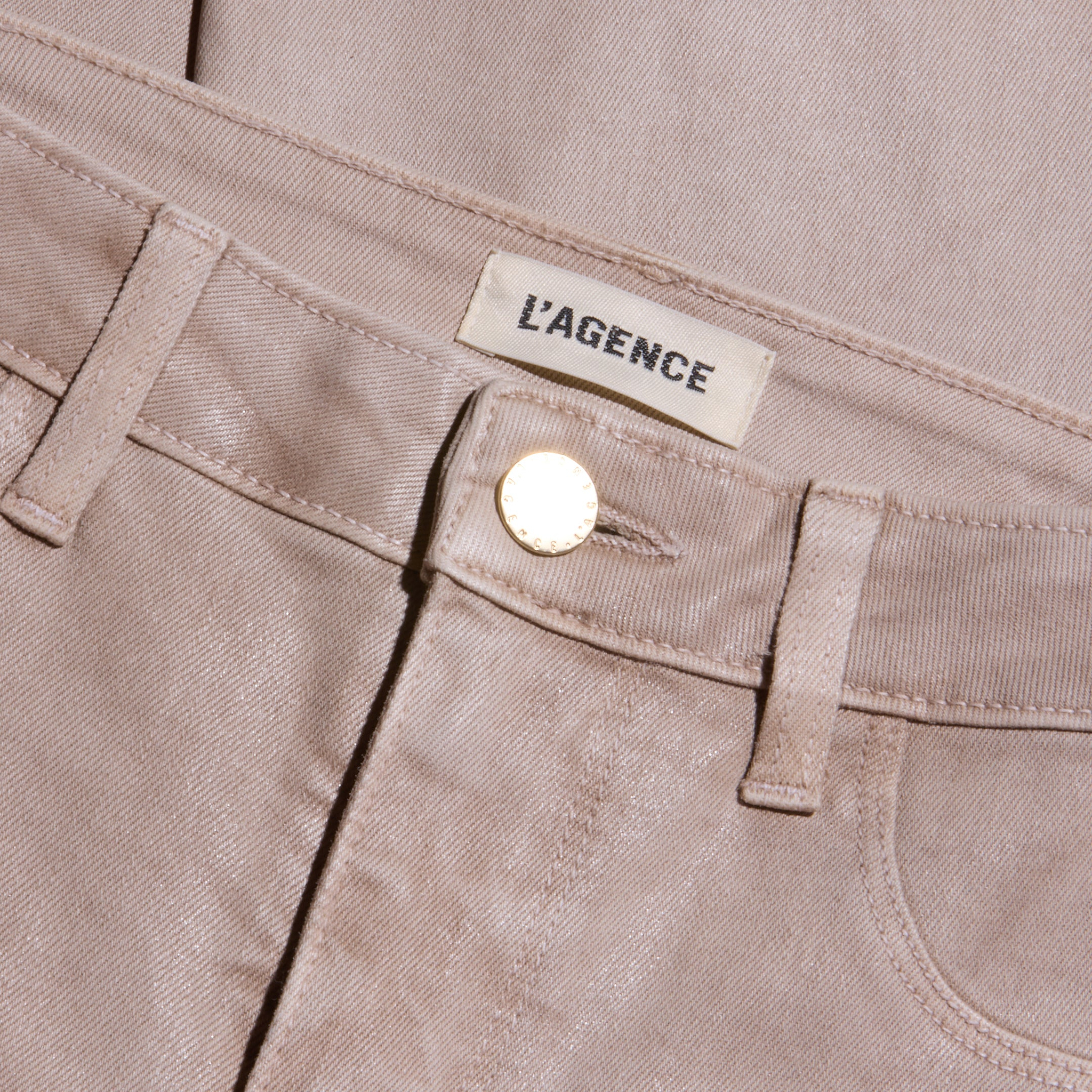 Close-up of the Kendra Coated Cropped Flare Jean in light beige, featuring a gold button and a fabric label above the waistband reading LAGENCE. The coated denim’s texture and detailed stitching are visible.