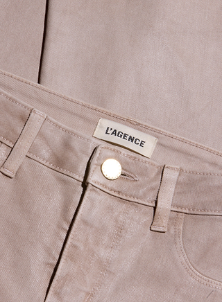Close-up of the Kendra Coated Cropped Flare Jean in beige, featuring a gold button and a label reading L’AGENCE above the waistband. The fabric showcases a slightly shiny, coated denim texture and the neatly folded jeans highlight their cropped flare silhouette.