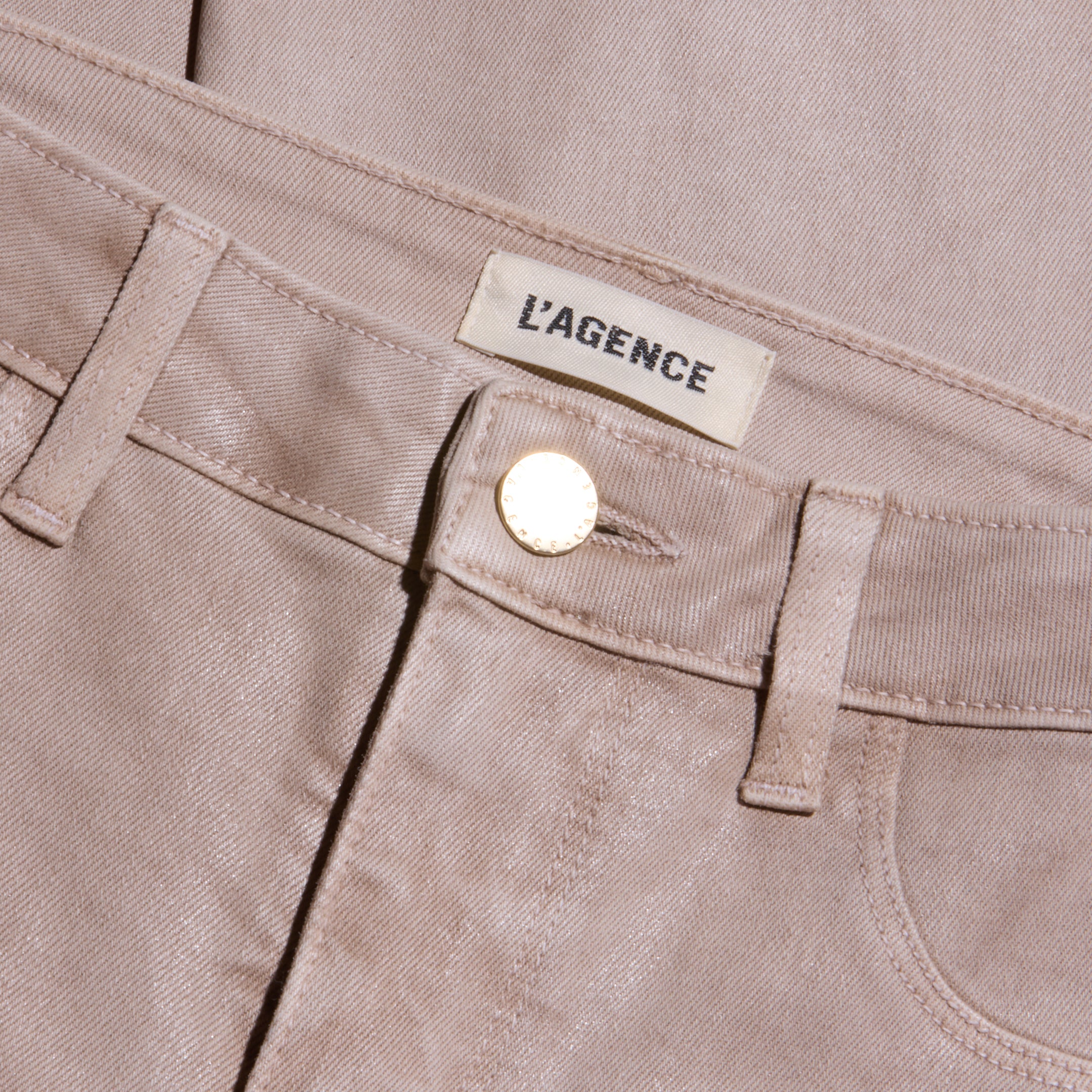 Close-up of the Kendra Coated Cropped Flare Jean in light beige, featuring a gold button and a fabric label above the waistband reading LAGENCE. The coated denim’s texture and detailed stitching are visible.