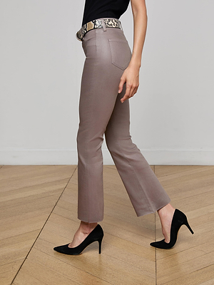 A person wearing L'AGENCE Kendra Coated Cropped Flare Jean in taupe, a black top, snake-print belt, and black high heels stands on a wooden floor against a plain light-colored wall.