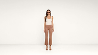 A woman wearing sunglasses, a white tank top, brown high-waisted pants, and brown heeled sandals stands against a plain white background.