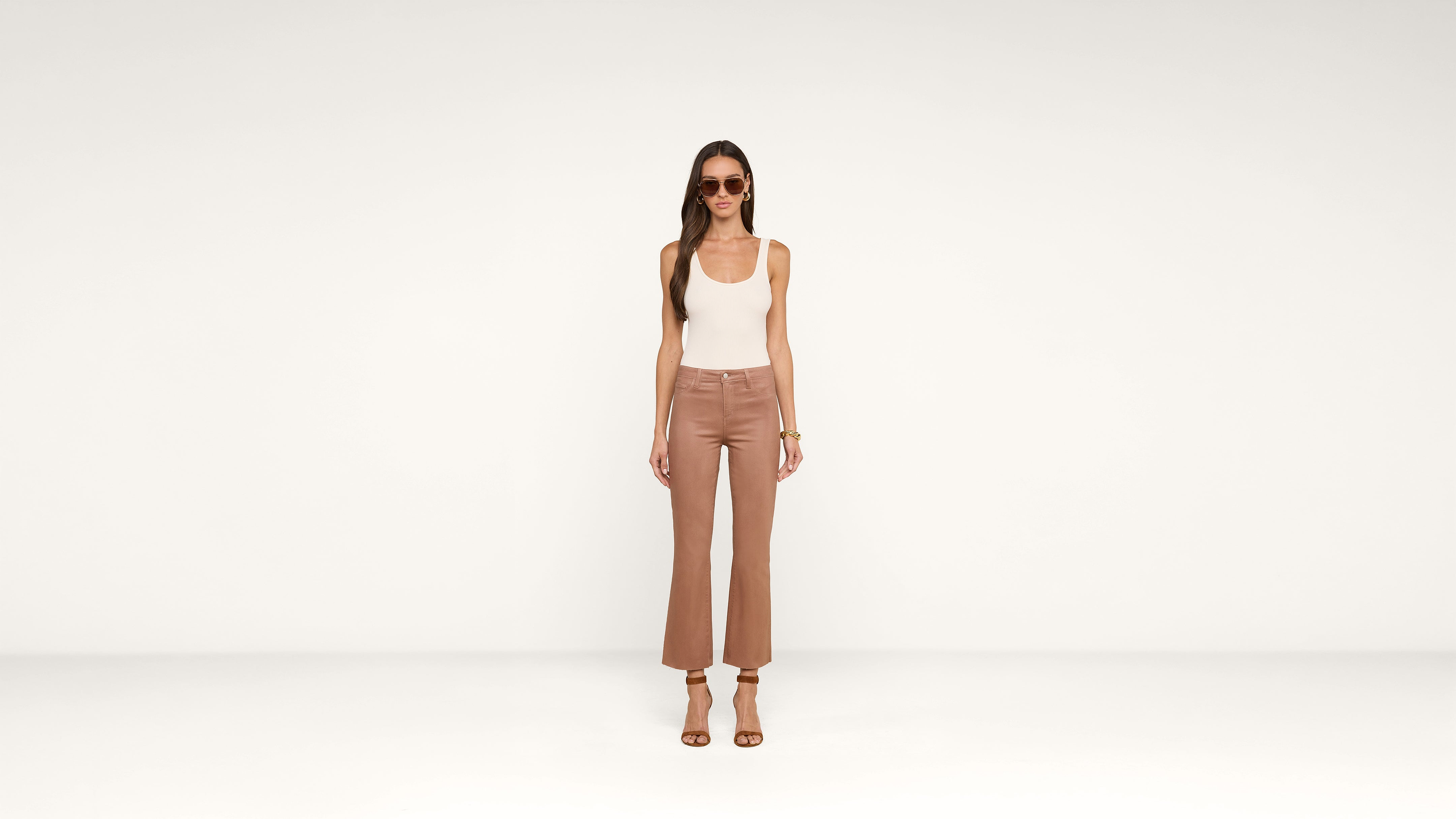 A woman wearing sunglasses, a white tank top, brown high-waisted pants, and brown heeled sandals stands against a plain white background.