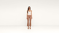 A woman stands in a minimalist setting wearing sunglasses, a white tank top, high-waisted brown pants, and heeled sandals, embodying effortless chic.