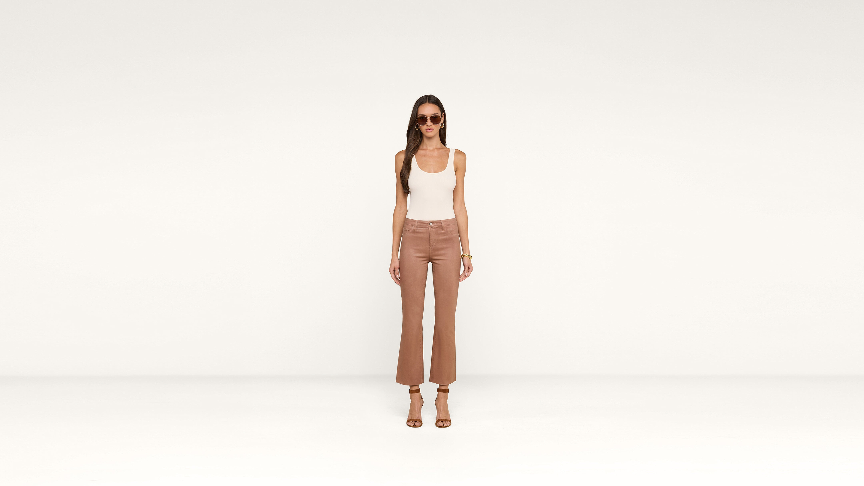 A woman stands in a minimalist setting wearing sunglasses, a white tank top, high-waisted brown pants, and heeled sandals, embodying effortless chic.