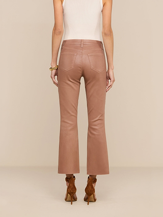 A woman stands facing away, wearing a white sleeveless top, the Petite Kendra Coated Jean in light brown high rise flared style, brown heels, and a gold bracelet, against a beige background.
