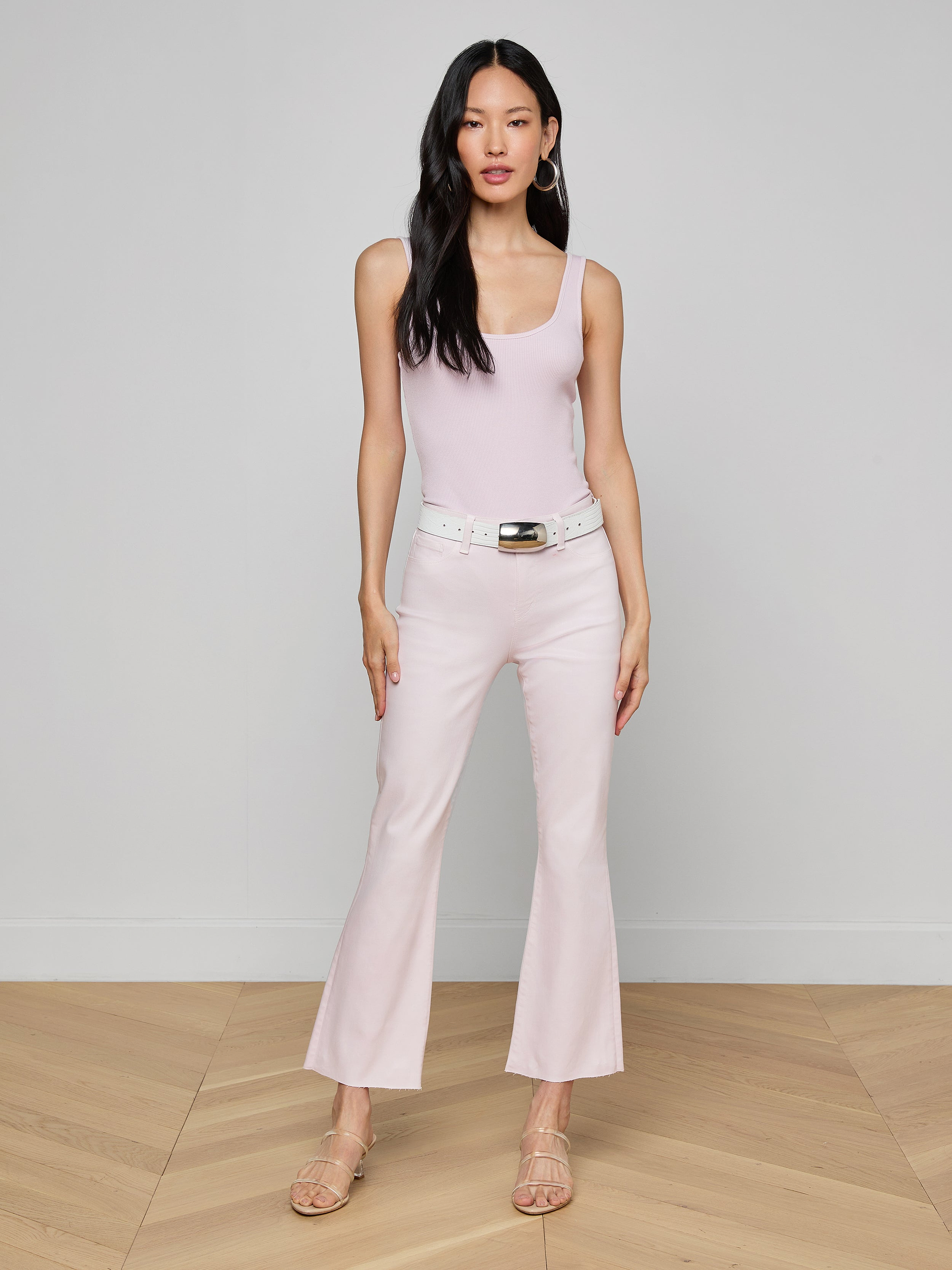 A woman with long dark hair and a neutral expression stands against a plain background, wearing a light pink sleeveless top paired with the Kendra Coated Cropped Flare Jean in a matching shade, styled with a white belt and beige heeled sandals.