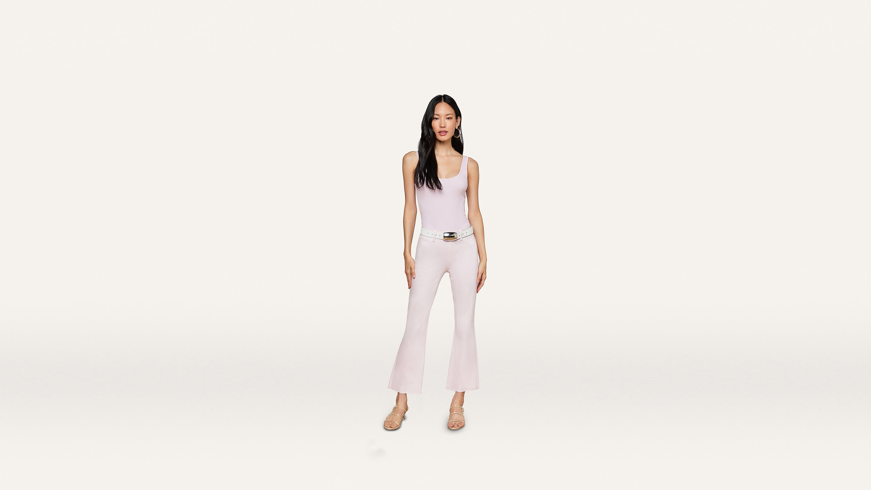 A woman stands facing forward, wearing a light pink sleeveless top and matching flared pants with a stylish white belt and beige heels, set against a minimalist white background.