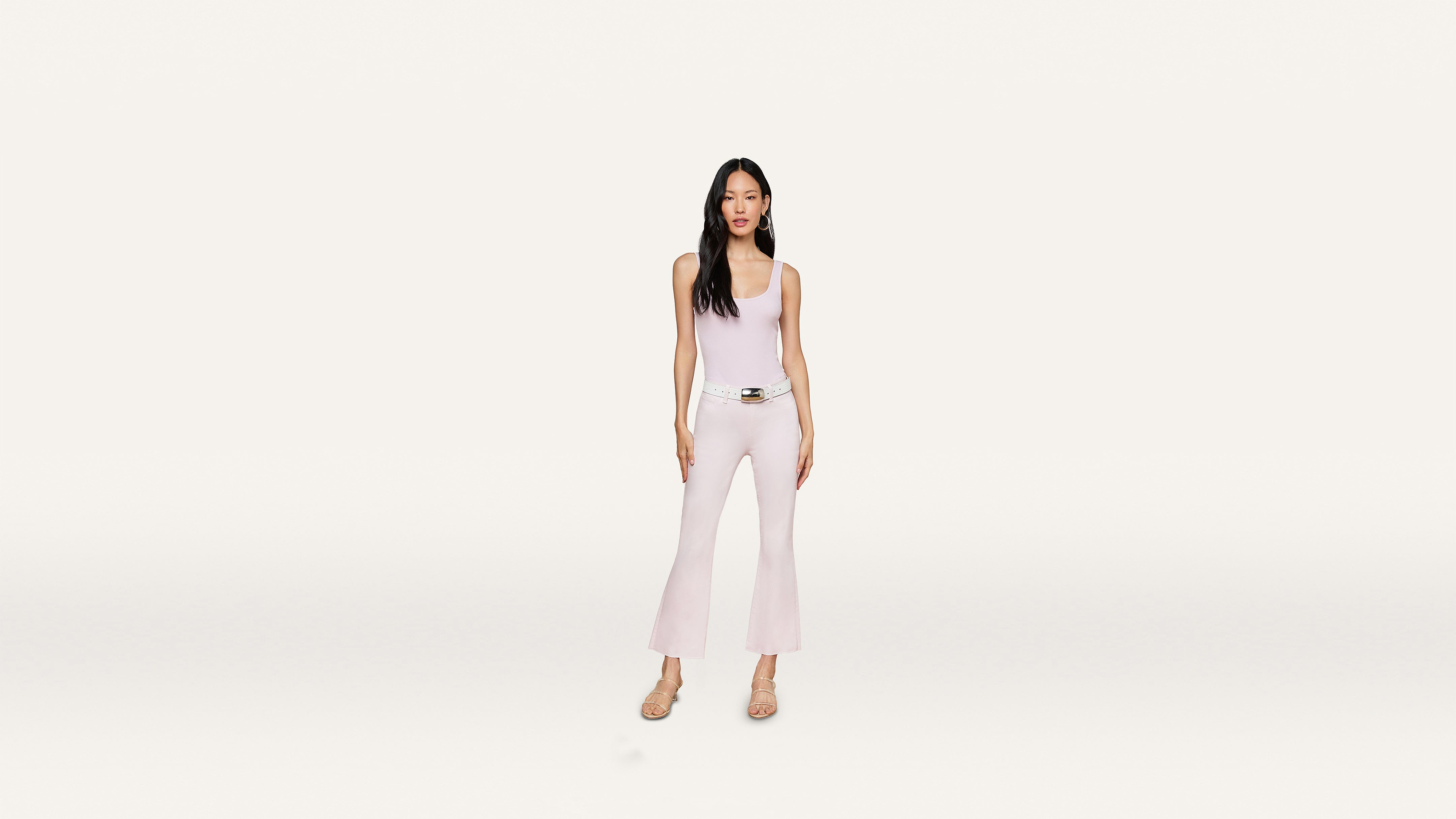 A woman stands facing forward, wearing a light pink sleeveless top and matching flared pants with a stylish white belt and beige heels, set against a minimalist white background.