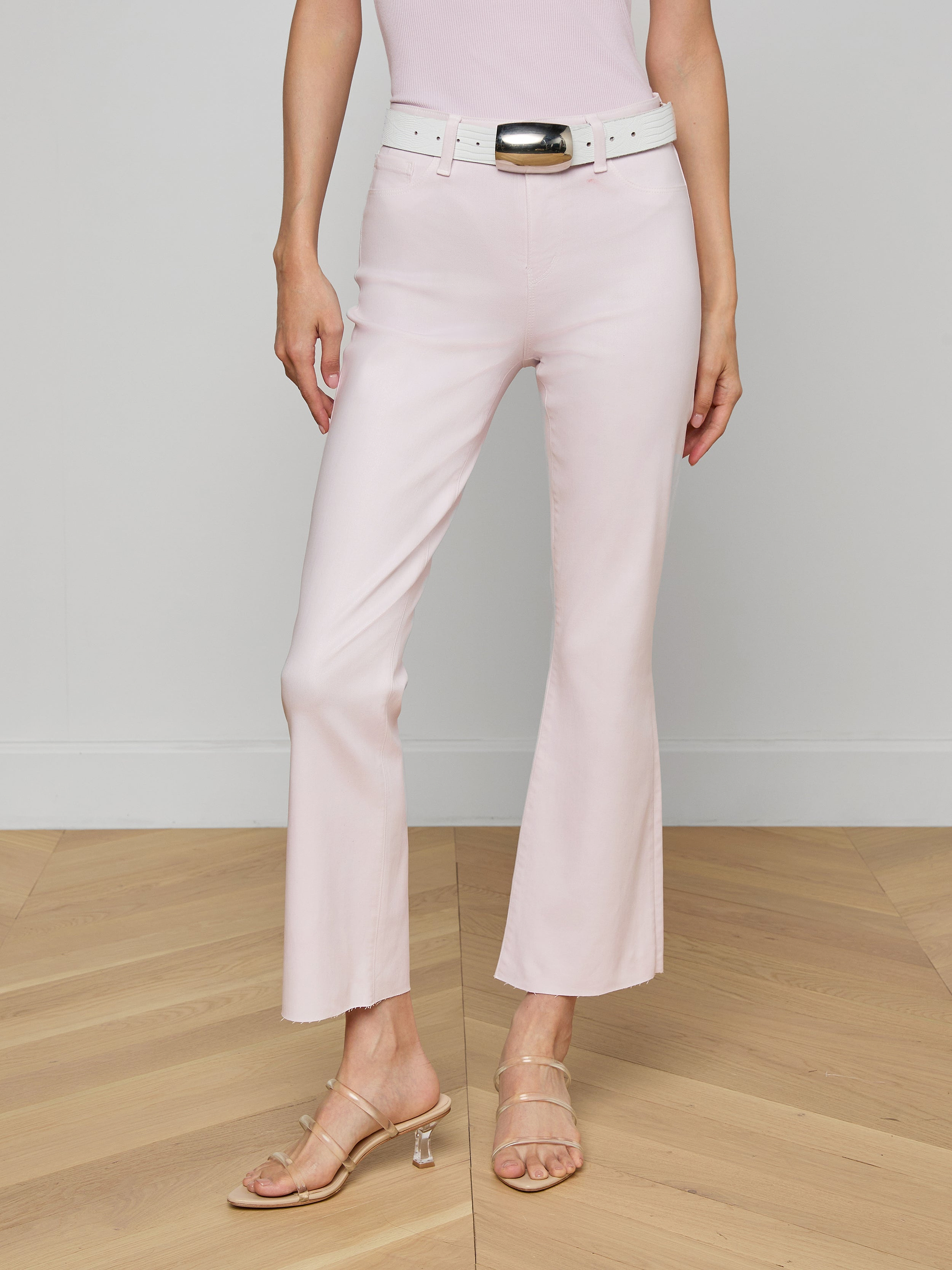 A person is wearing the Kendra Coated Cropped Flare Jean in light pink with a matching belt and beige heeled sandals, standing against a plain white background. Their upper body is partially out of frame.