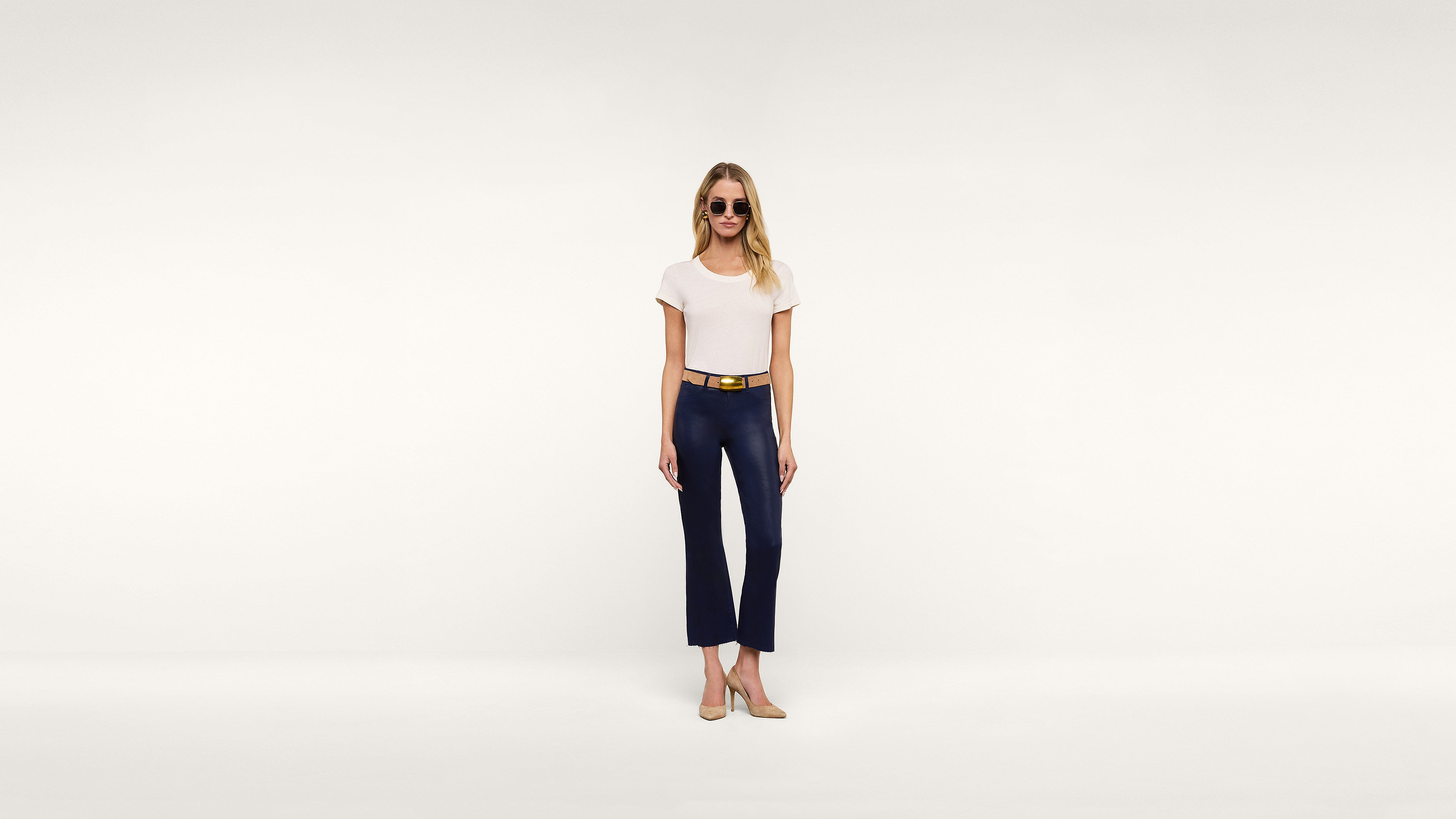 A woman wearing sunglasses, a white t-shirt, navy pants, a belt, and beige heels stands confidently against a plain white background.