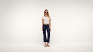 A woman wearing sunglasses, a white T-shirt, navy cropped pants, a tan belt, and beige shoes stands against a plain white background.