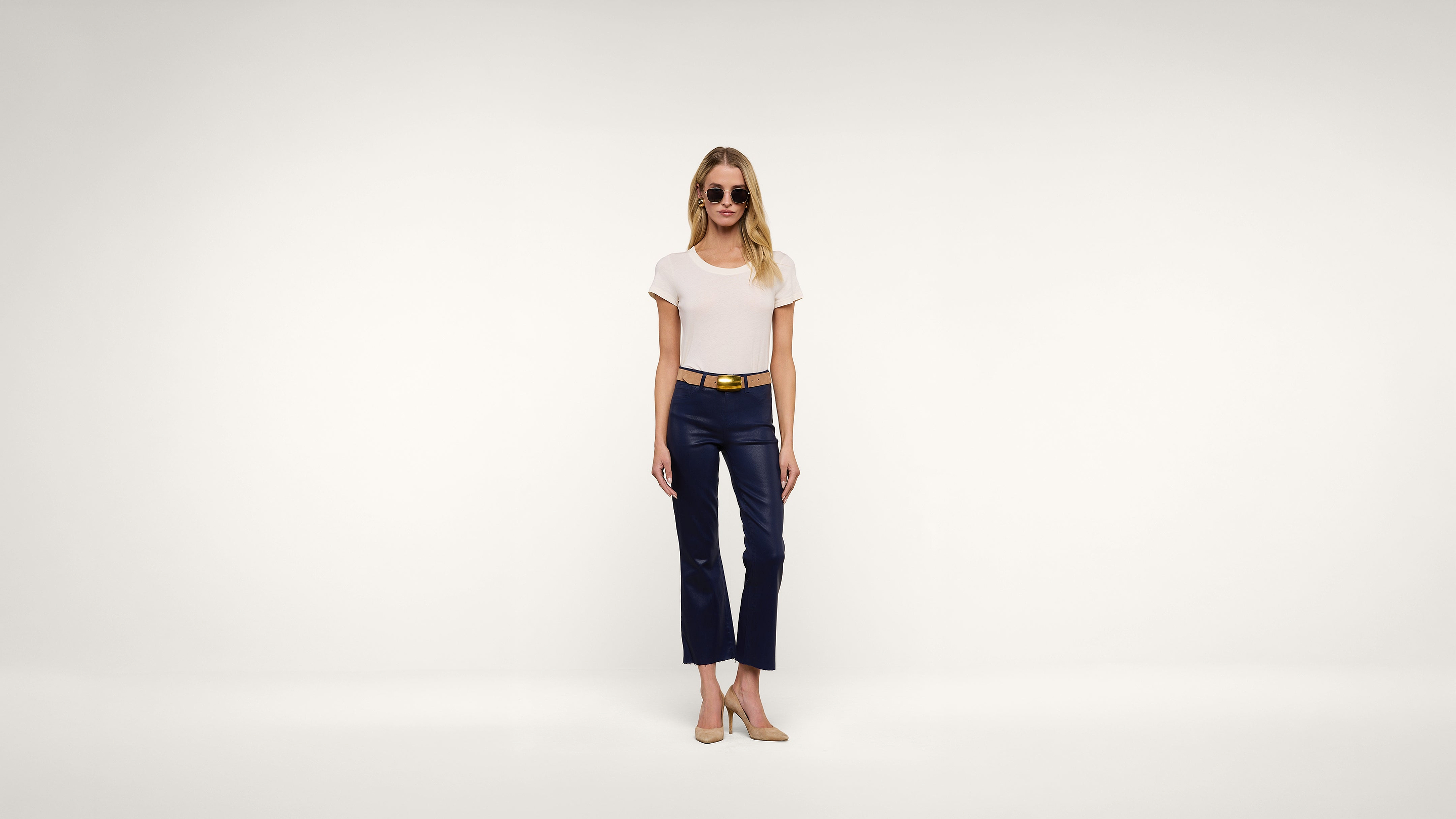 A woman wearing sunglasses, a white T-shirt, navy cropped pants, a tan belt, and beige shoes stands against a plain white background.