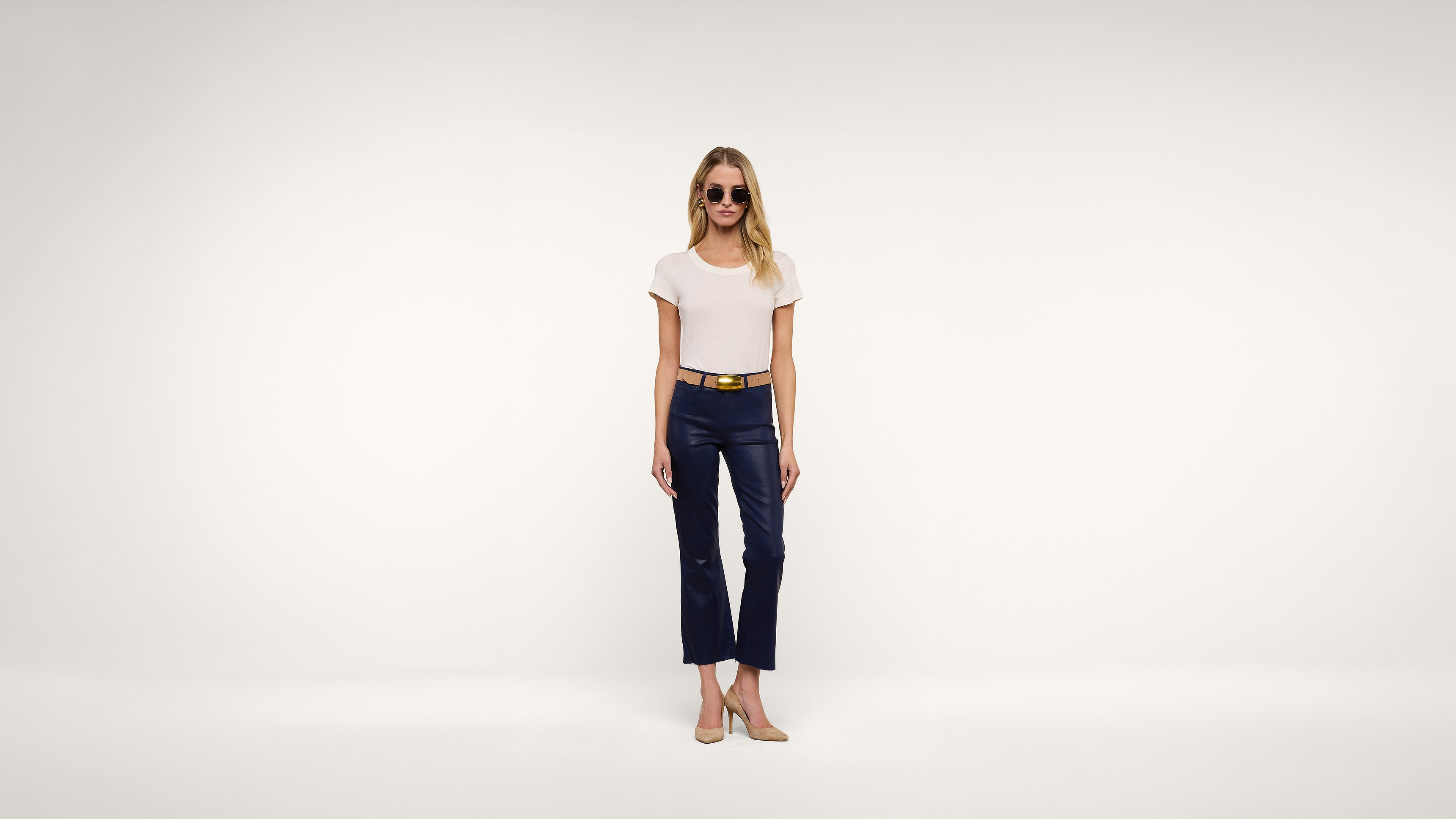 A woman wearing sunglasses, a white T-shirt, navy cropped pants, a tan belt, and beige shoes stands against a plain white background.