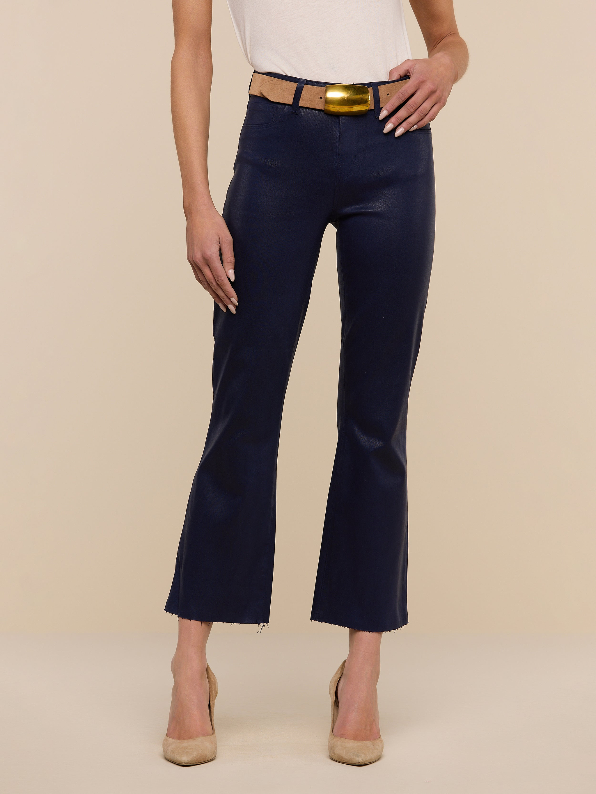 A person stands against a plain white background wearing a sleeveless beige top, a tan belt, beige heels, and the Kendra Coated Cropped Flare Jean in dark blue.