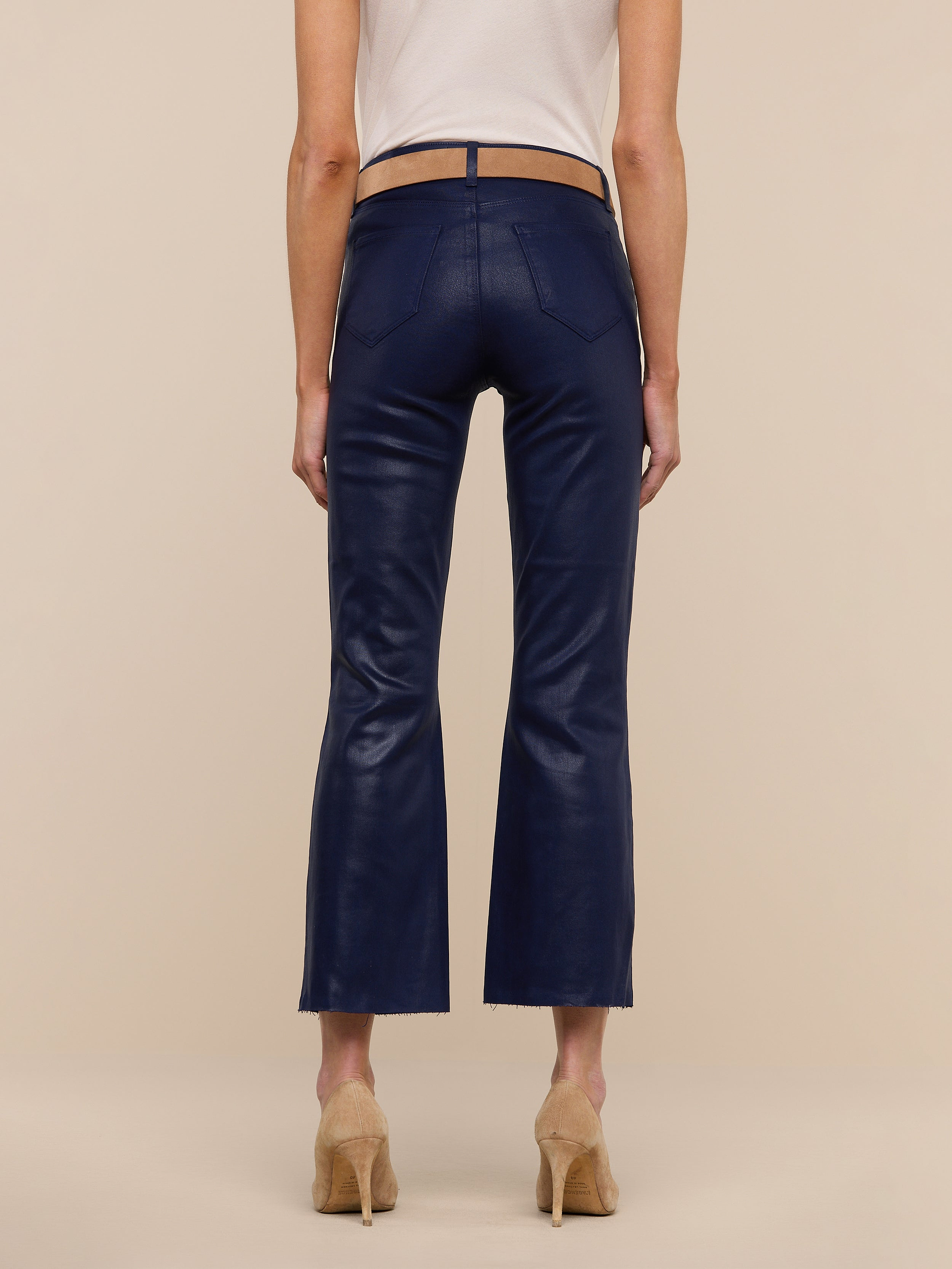 A person wearing the Kendra Coated Cropped Flare Jean in dark blue, paired with a beige top and tan high-heeled shoes, is standing facing away from the camera against a plain white background.
