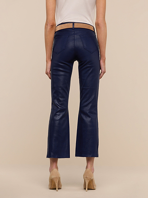 A person stands facing away, wearing the Kendra Coated Cropped Flare Jean in navy blue with a raw hem, paired with a white sleeveless top and beige heeled shoes against a plain beige background.