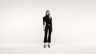 A woman wearing a black top, black pants, and black heels stands against a plain white background, facing forward with her arms relaxed by her sides.