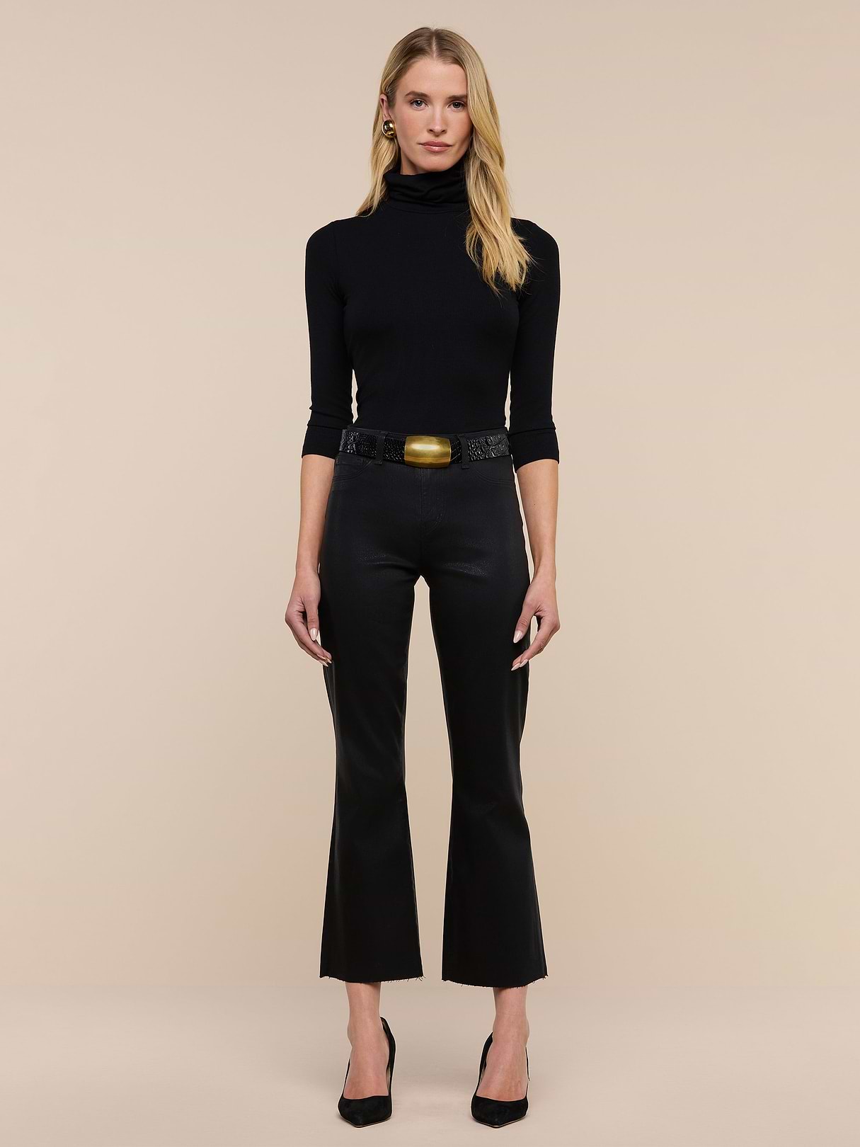A woman with long blonde hair stands against a beige background, wearing a black turtleneck, Kendra Coated Cropped Flare Jean, a textured belt with a gold buckle, black heels, and gold earrings. She faces the camera with a neutral expression.