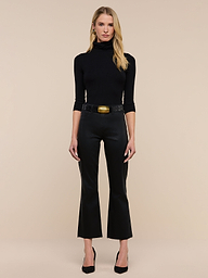 A woman with long blonde hair and a neutral expression stands against a plain white background, wearing black heels, a black turtleneck, a wide belt with a large gold buckle, and the Kendra Coated Cropped Flare Jean.