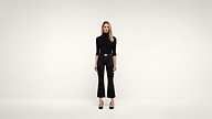 A woman wearing a black turtleneck, black pants, and black heels stands against a plain white background, facing forward with a neutral expression.