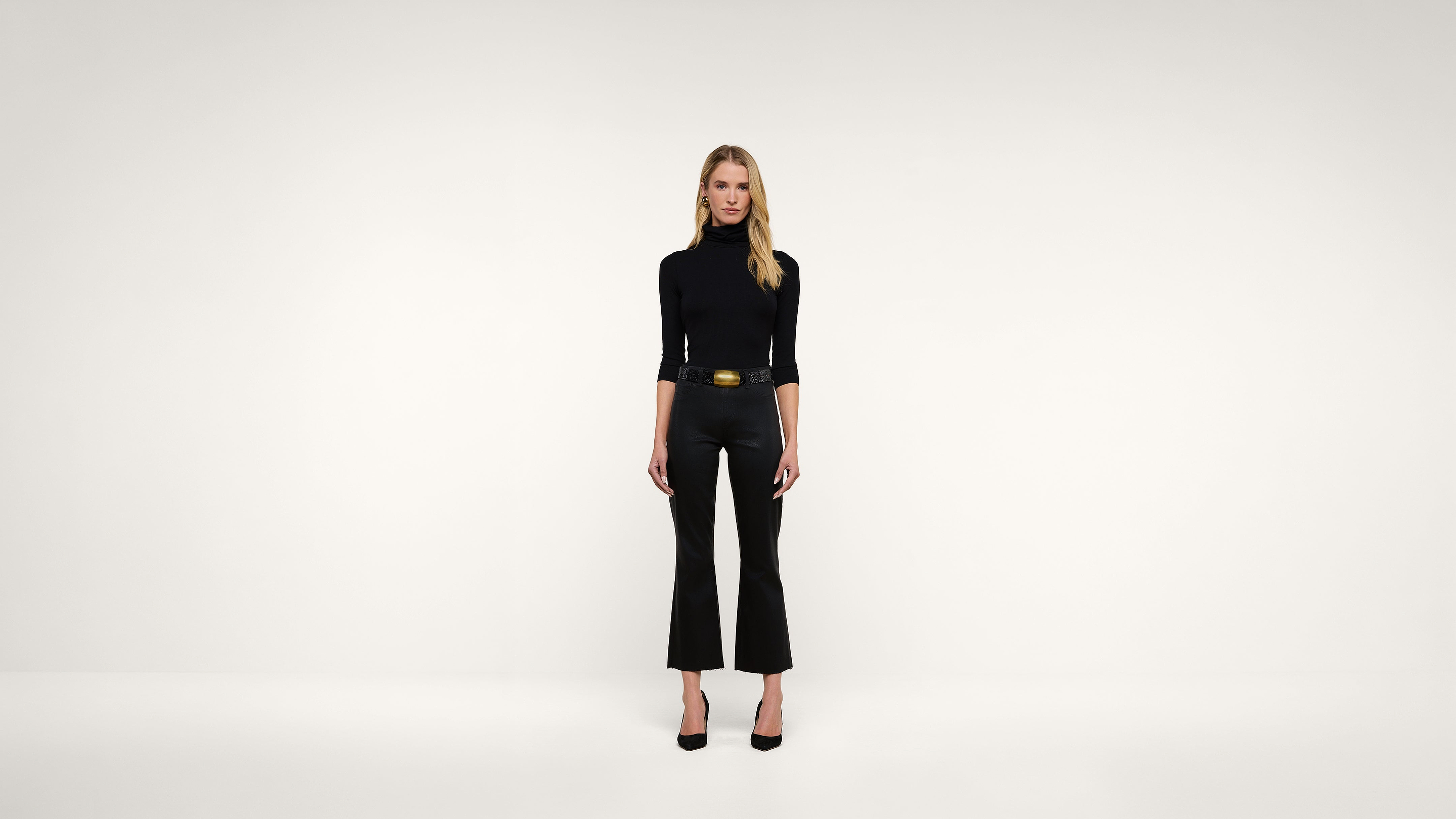 A woman wearing a black turtleneck, black pants, and black heels stands against a plain white background, facing forward with a neutral expression.
