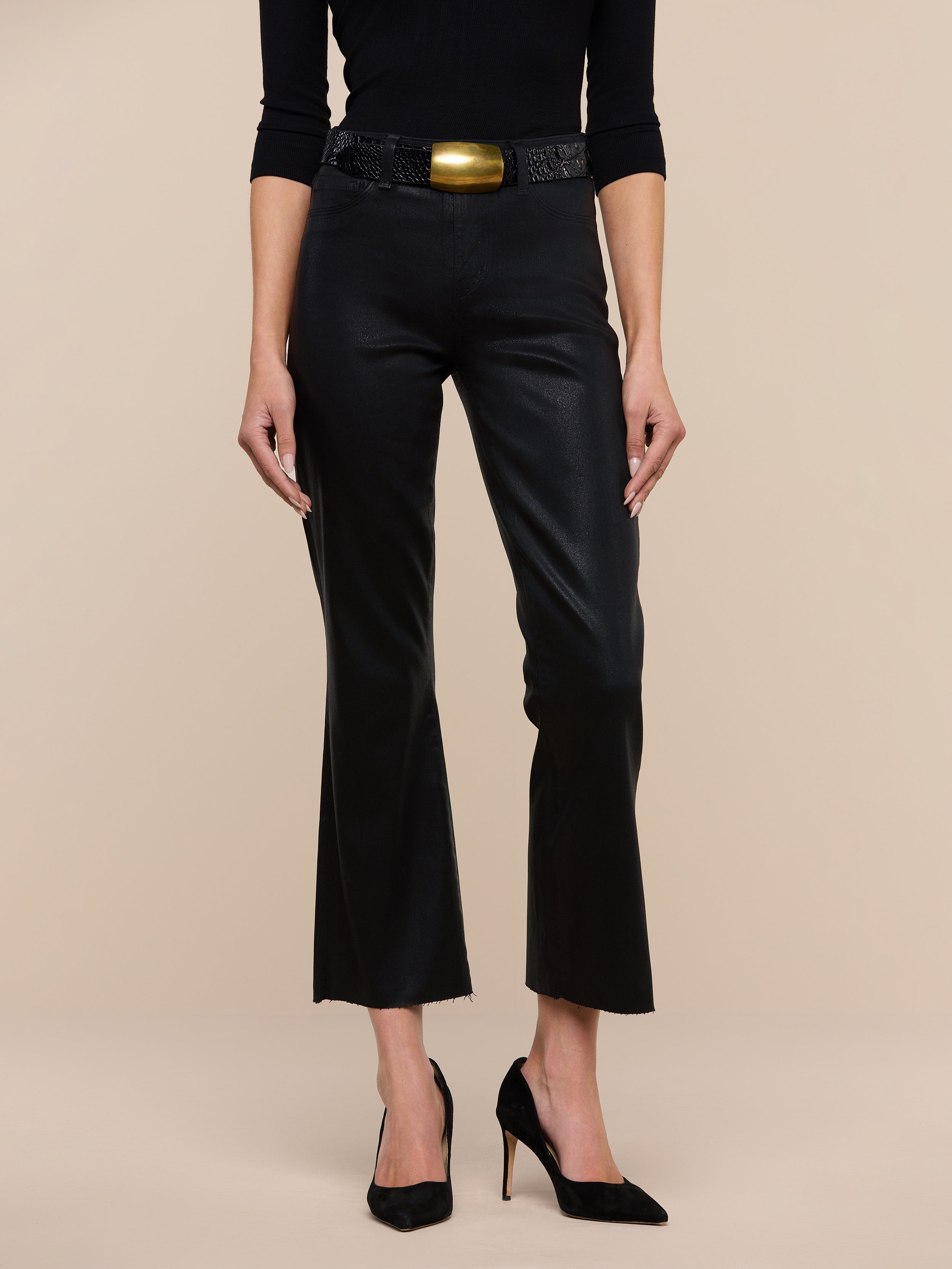 A woman is shown wearing the Kendra Coated Cropped Flare Jean in black, styled with a matching long-sleeve top, high heels, and a statement belt with a large gold buckle. She stands against a plain white background, with her head and feet cropped out of the image.