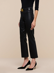 A person stands against a plain white background, wearing the Kendra Coated Cropped Flare Jean with a high-rise fit, styled with a black belt featuring a gold buckle, a long-sleeve black top, and black high-heeled shoes.
