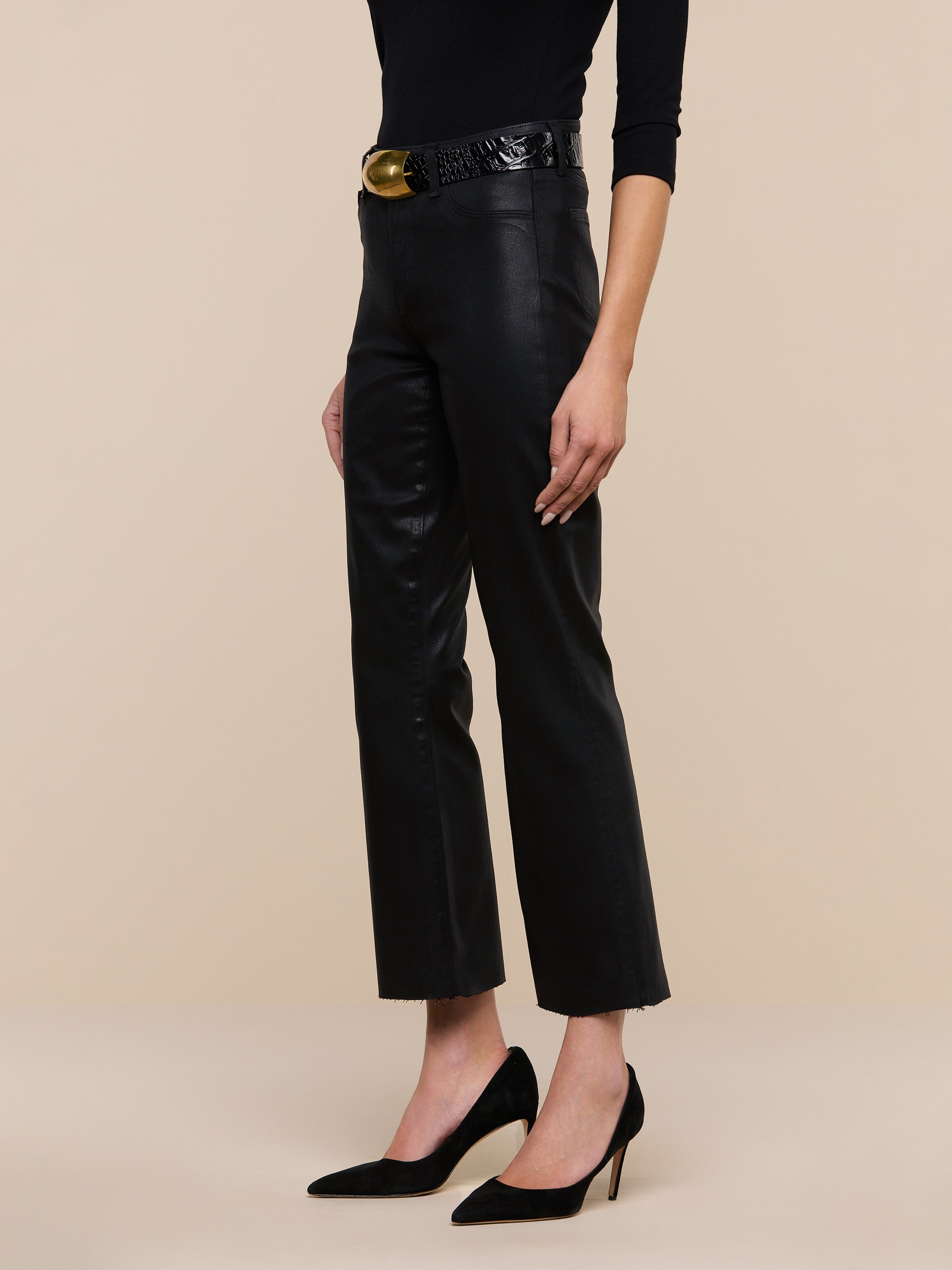A person stands against a plain white background, wearing the Kendra Coated Cropped Flare Jean with a high-rise fit, styled with a black belt featuring a gold buckle, a long-sleeve black top, and black high-heeled shoes.