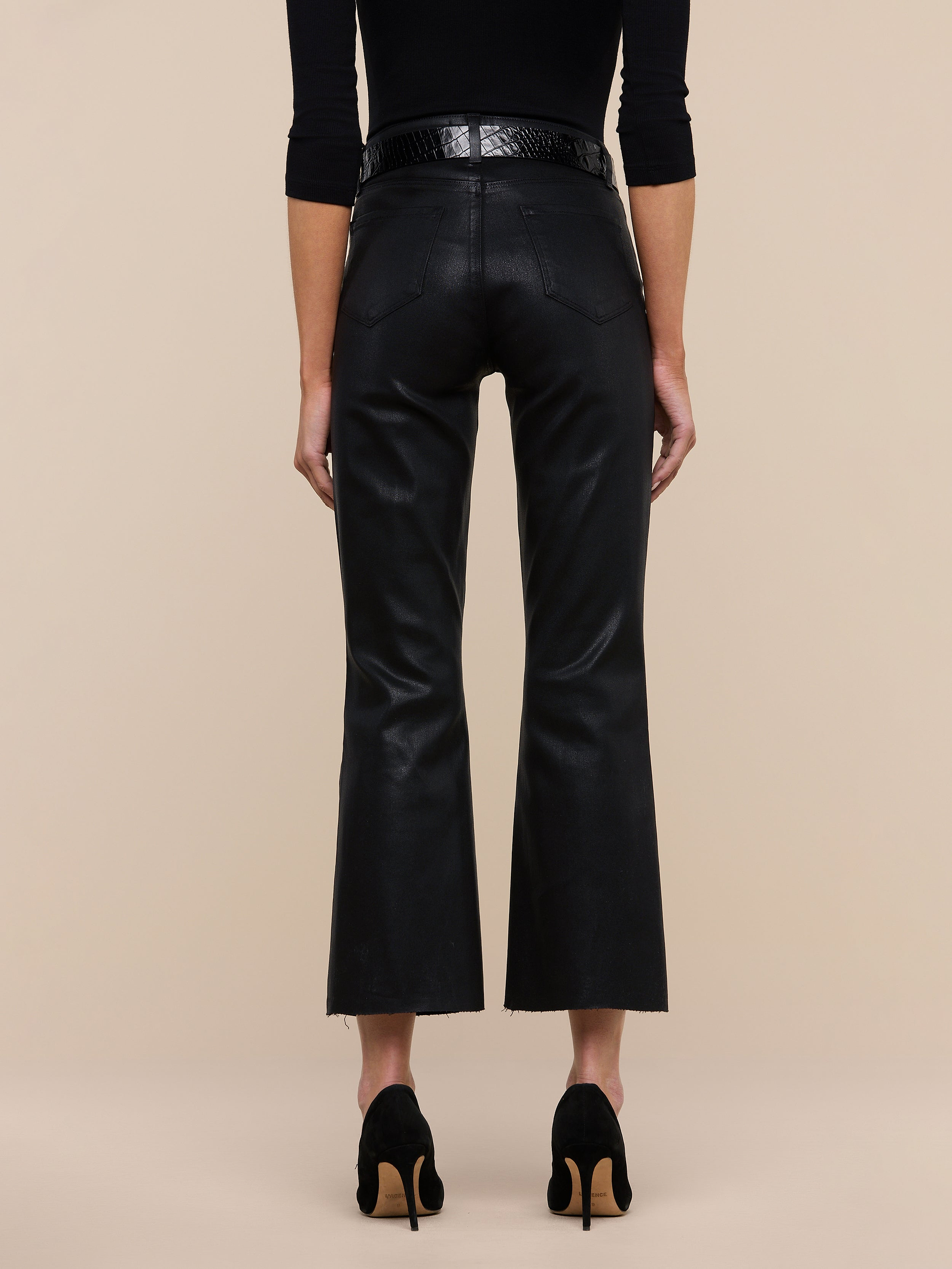 A person stands facing away against a plain white background, wearing the Kendra Coated Cropped Flare Jean in high-rise black coated denim, styled with a black belt, a black long-sleeve top, and black heeled shoes.
