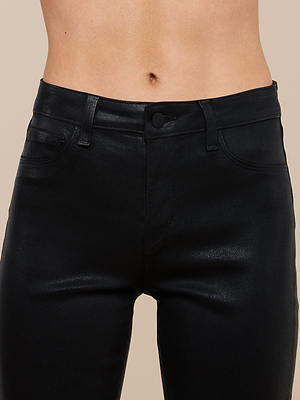 Close-up of a persons midsection wearing the Kendra Coated Cropped Flare Jean in shiny black denim with a button and belt loops, set against a neutral beige background.