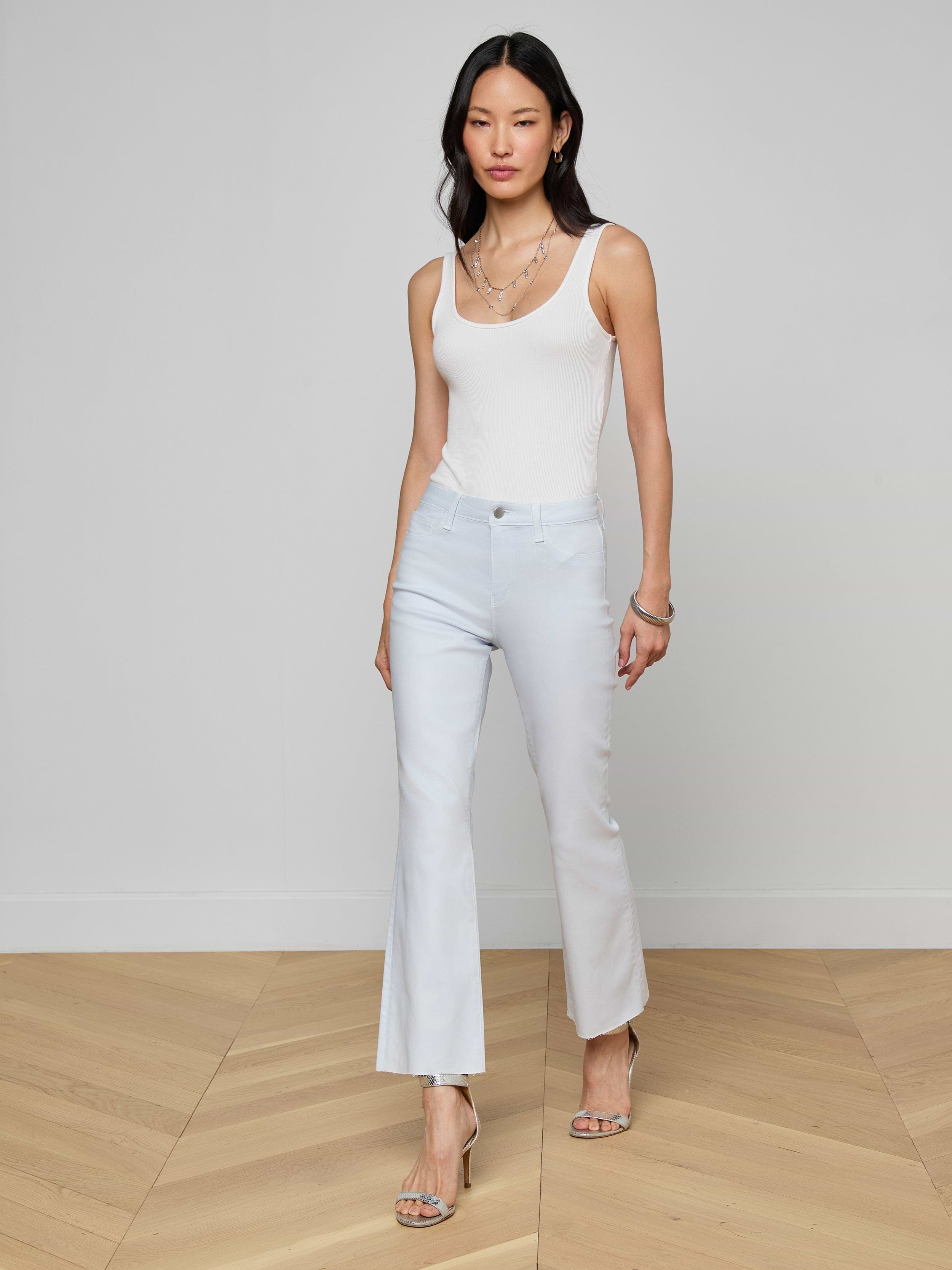 A woman with long dark hair stands indoors on a wooden floor, wearing a white sleeveless top and Kendra Coated Cropped Flare Jeans with heeled sandals, accessorized with layered necklaces and bracelets.