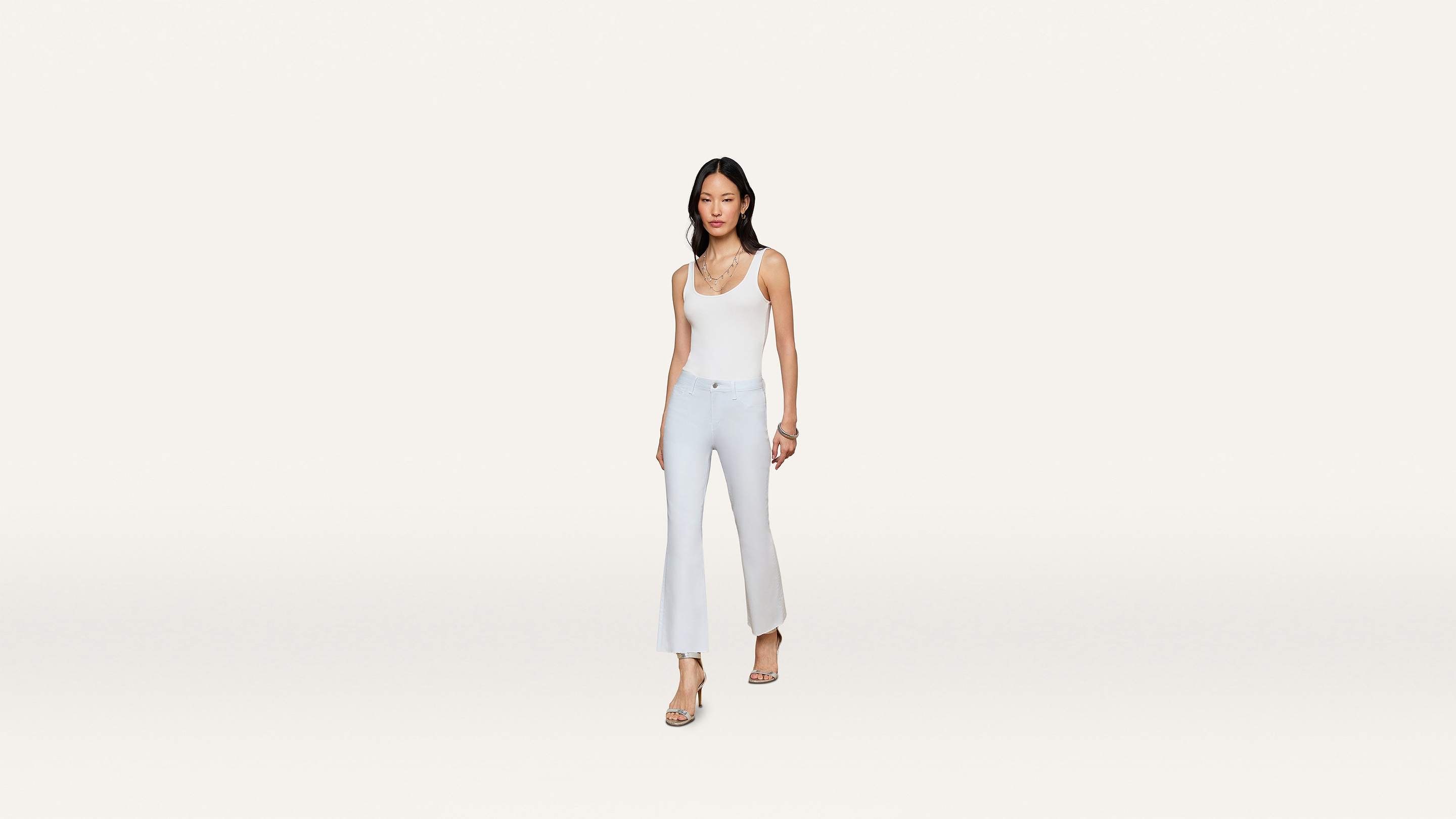 A woman in a white sleeveless top and light blue flared pants walks forward, wearing sandals and a delicate necklace, set against a plain white background.